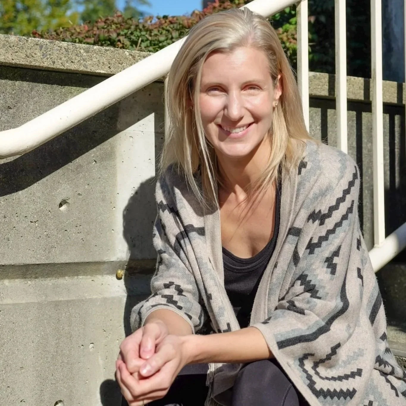 A smiling woman with blonde hair sitting outdoors on concrete steps, wearing a patterned cardigan and black top.