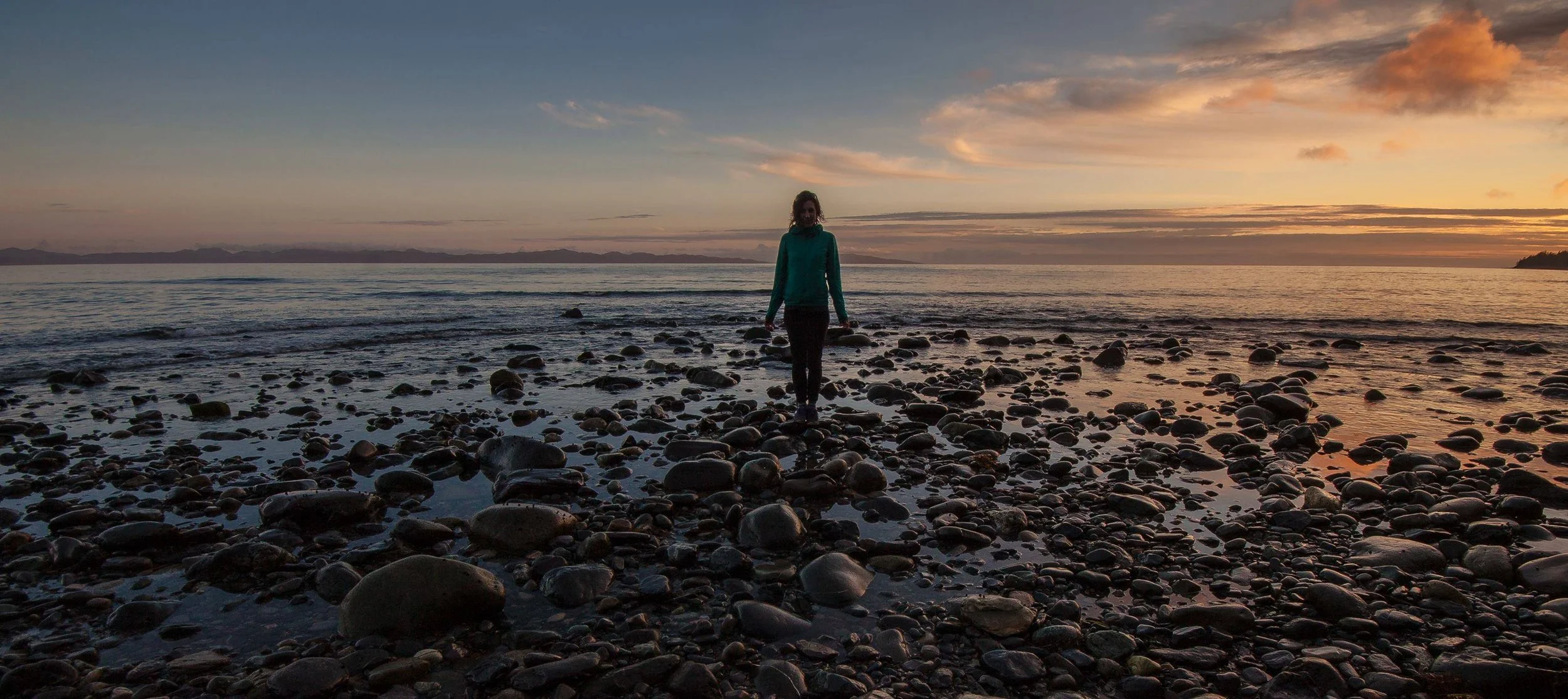 A person walking on a rocky beach at sunset with a colorful sky and calm ocean in the background.