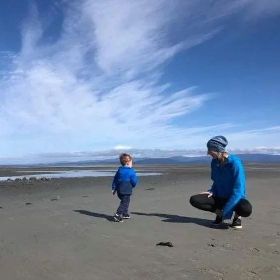 A woman and a young child on a sandy beach with a cloudy sky, ocean in the background, and mountains in the distance.