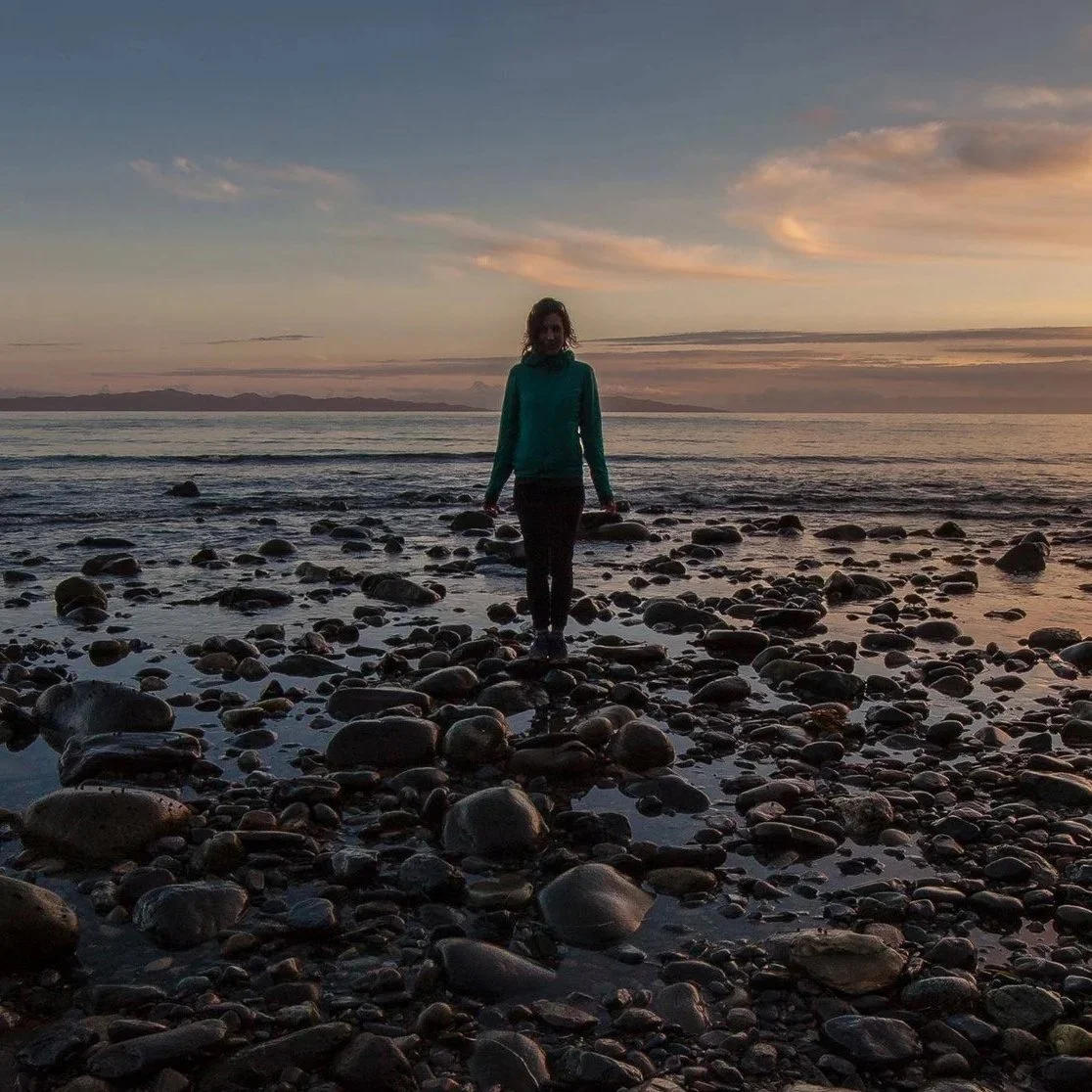 A woman standing on a rocky beach during sunset, with the ocean and mountains in the background.