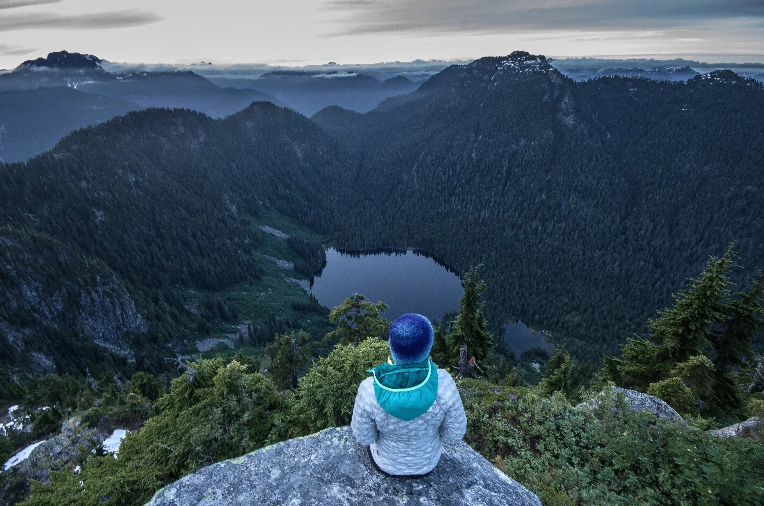 Person sitting on a rock overlooking a mountainous landscape with a lake in the valley below, surrounded by dense evergreen trees and distant mountain peaks under cloudy sky.