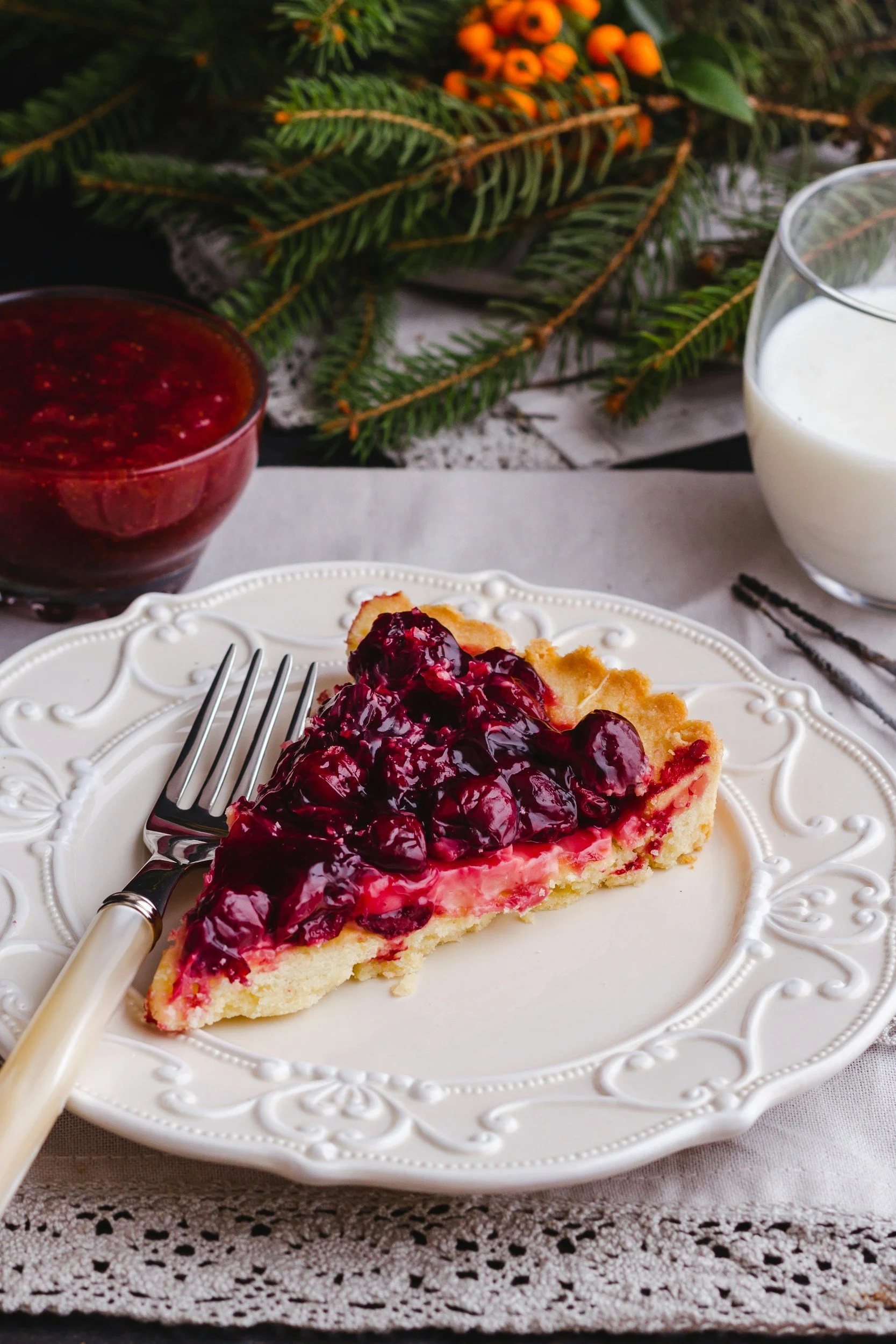 The photo features a slice of cherry tart served on an ornate white plate, accompanied by a glass of milk and a bowl of cherry sauce, with a festive greenery background.