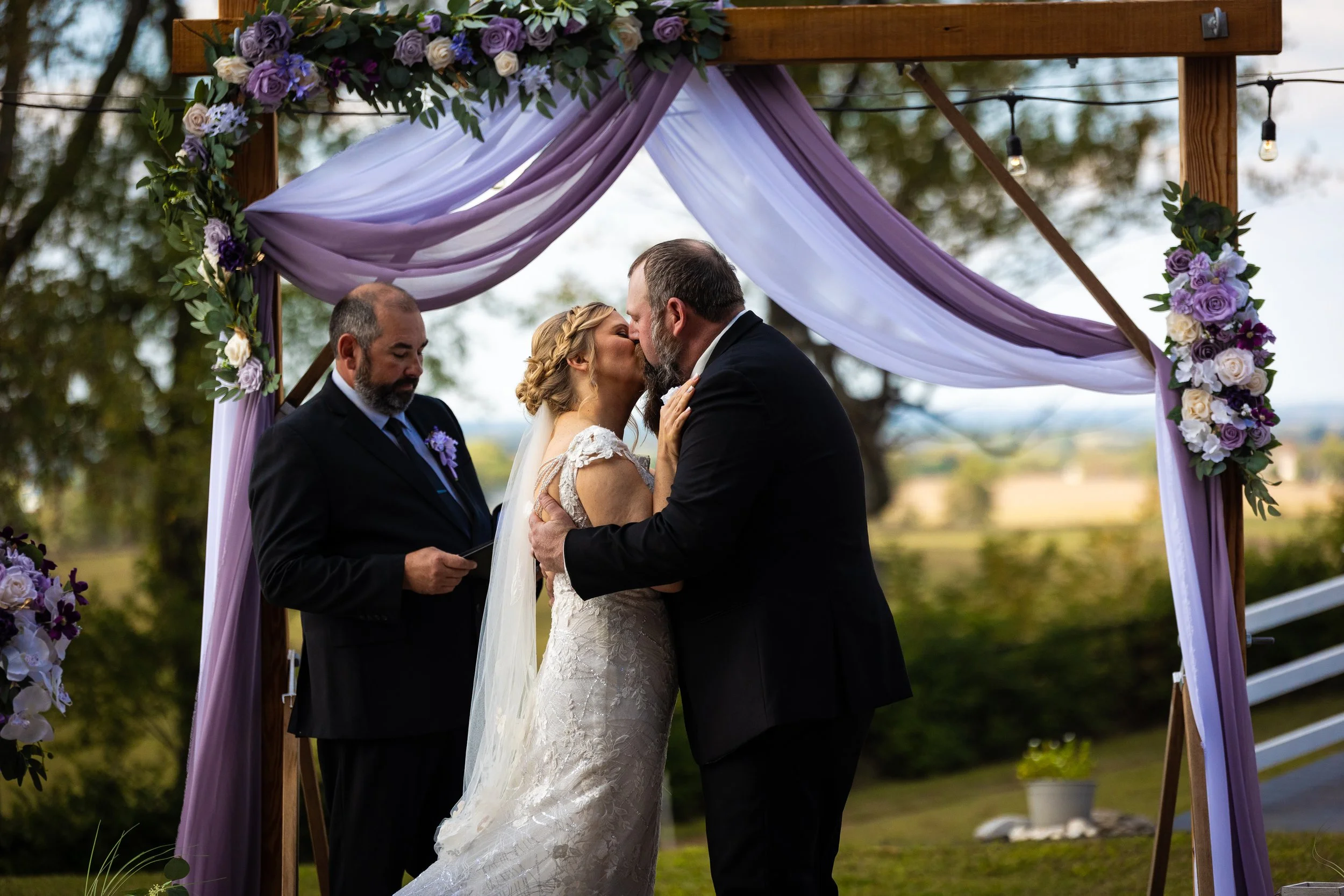 Bride and groom kissing under a wooden arch decorated with purple and white drapes and flowers, with officiant standing nearby during outdoor wedding ceremony.