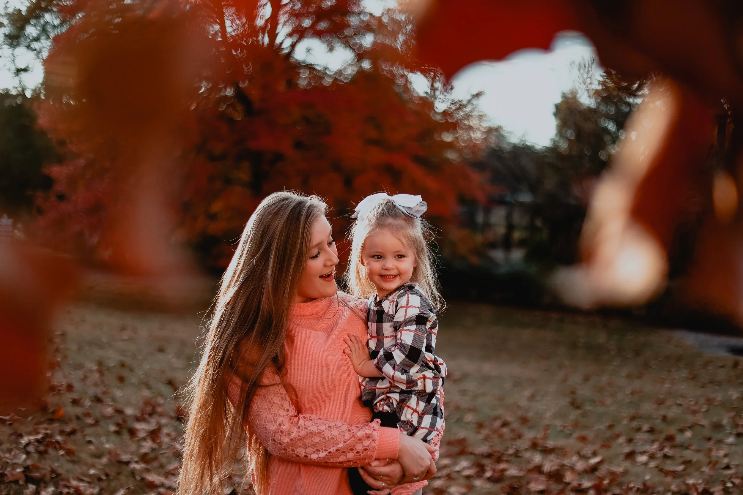 Woman in a pink sweater holding a young girl in a park with autumn leaves.
