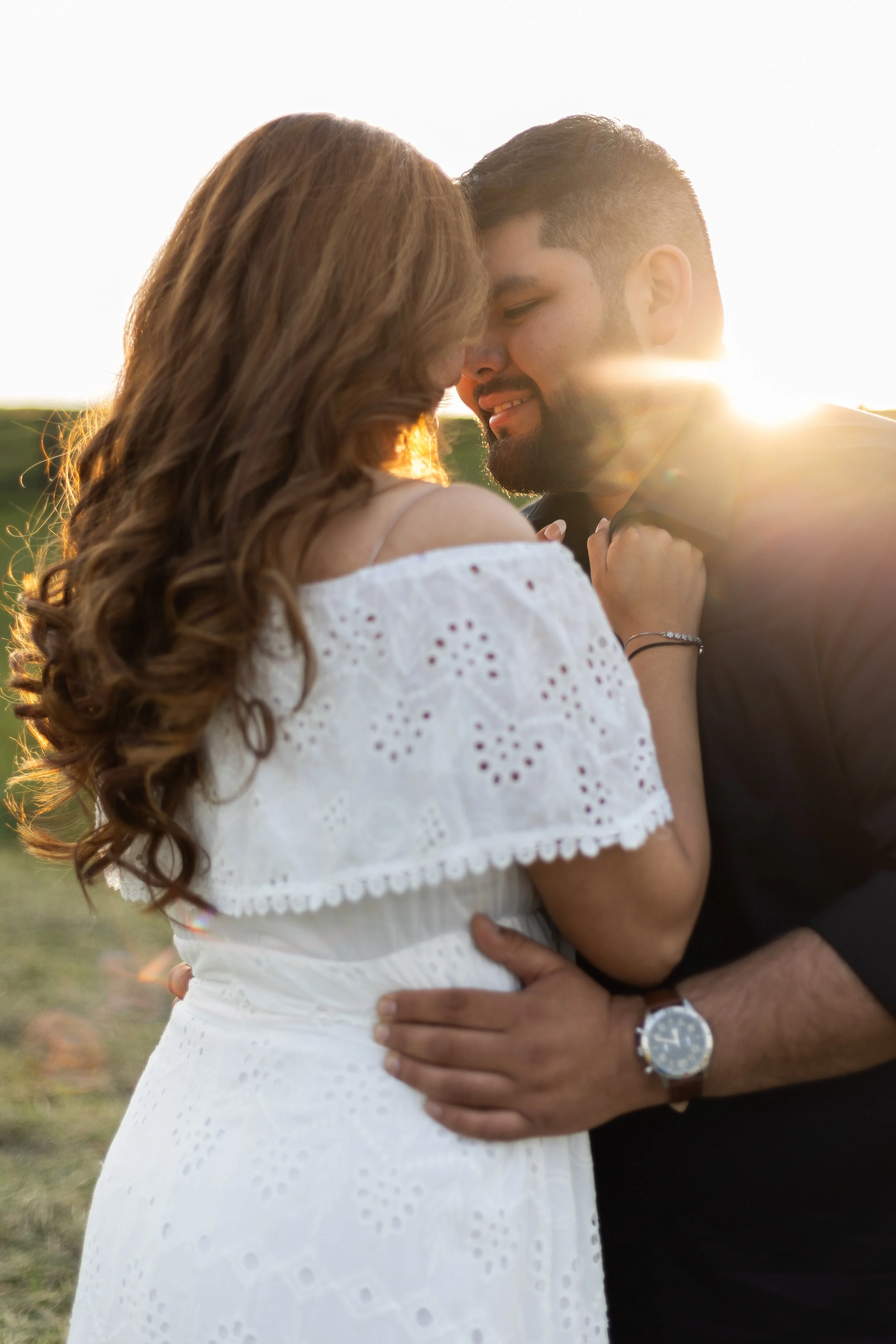 Couple embracing at sunset with lens flare, woman in white dress, man in black shirt.