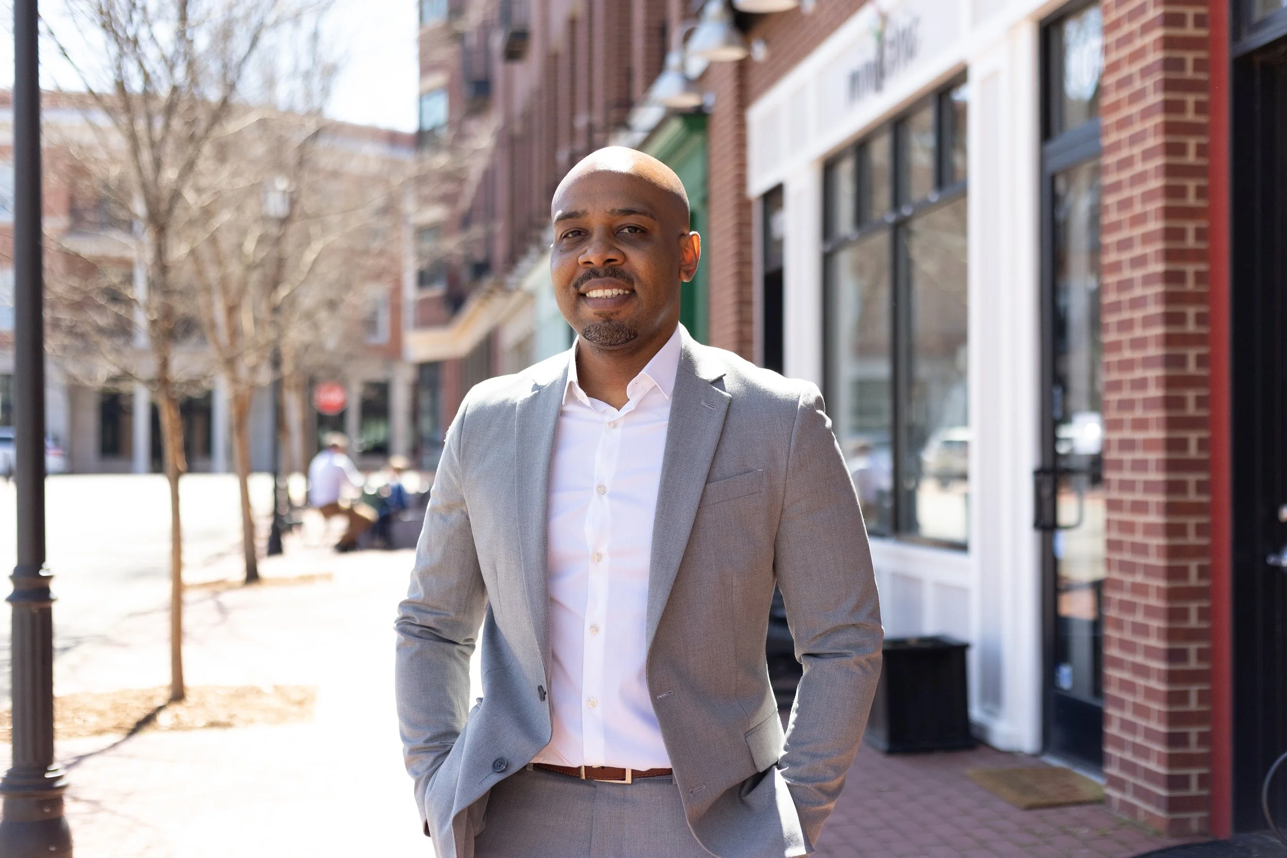 Man in a gray suit standing on a city sidewalk near a brick building, smiling.