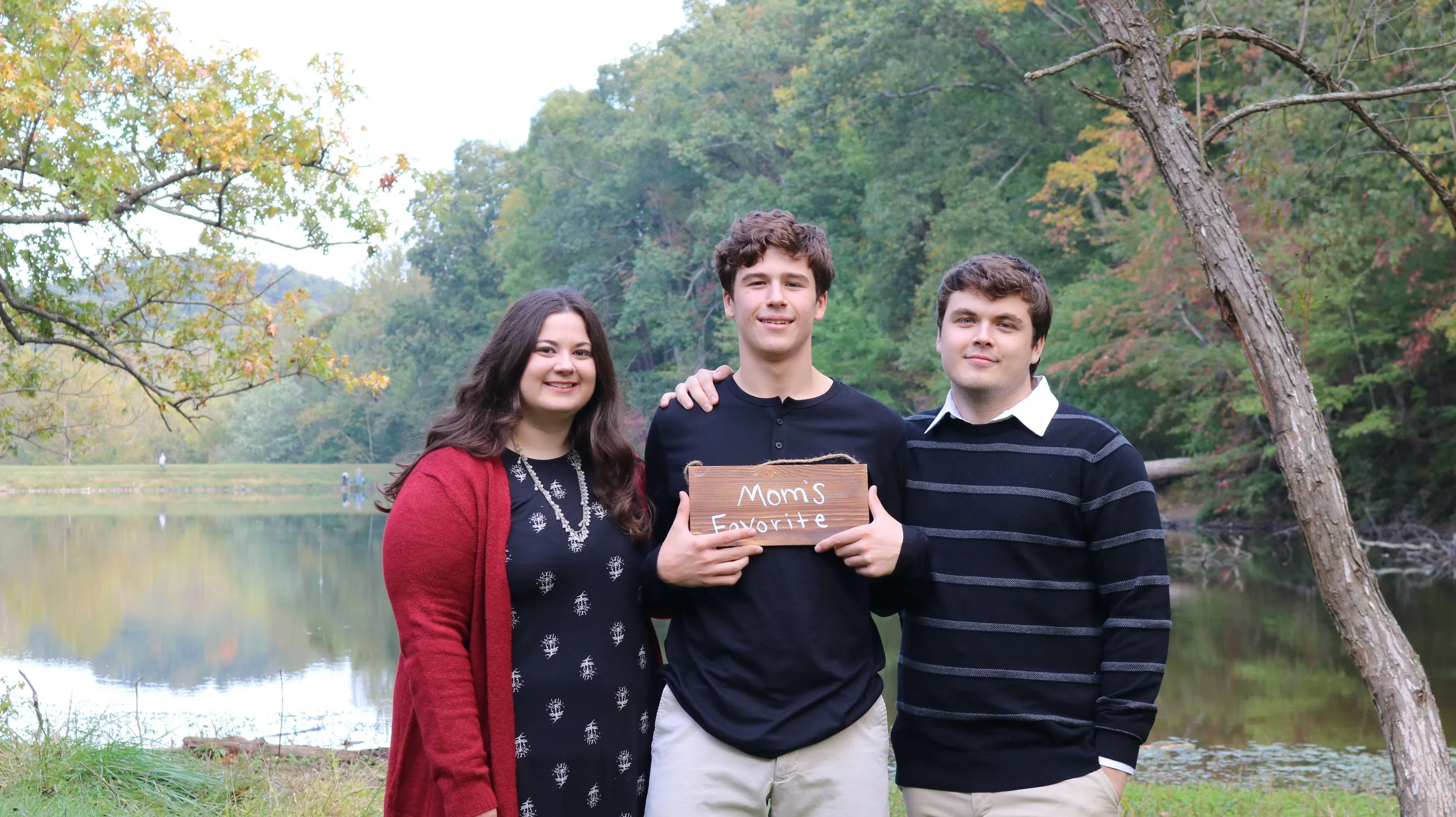 Three people outdoors by a lake with a sign reading "Mom's Favorite"; trees and autumn foliage in the background.