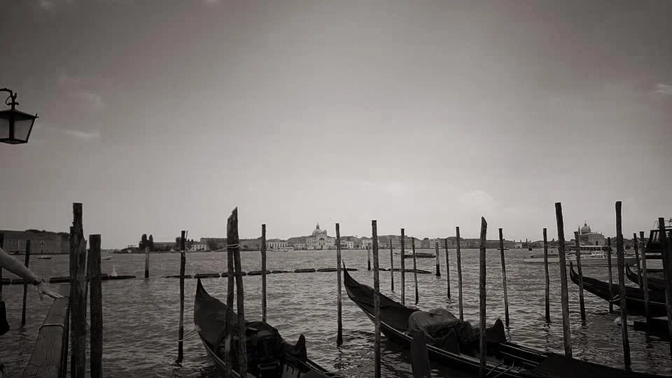 Black and white image of gondolas on a canal in Venice, Italy, with buildings in the background and wooden poles in the water.