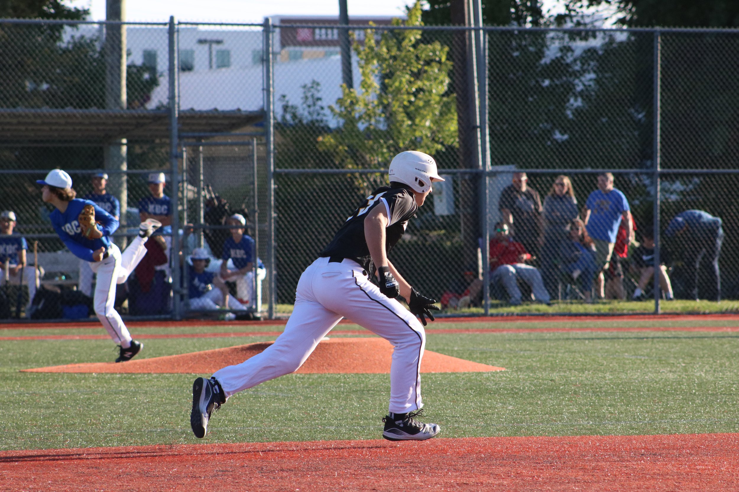 Baseball game with a player running towards first base and pitcher in mid-throw, with a dugout and spectators in the background.