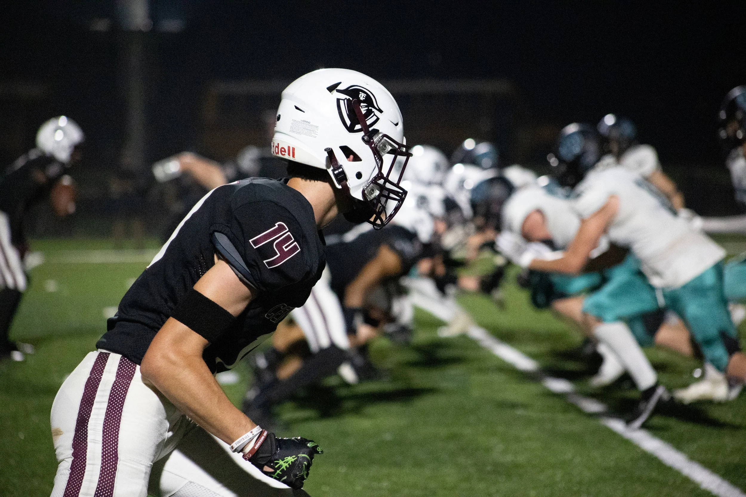 Football players on the field during a night game, with one in a black uniform and white helmet in the foreground and others lined up in the background facing off.
