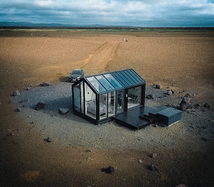 A modern glass cabin with a black frame in a desert landscape, featuring a car parked nearby.