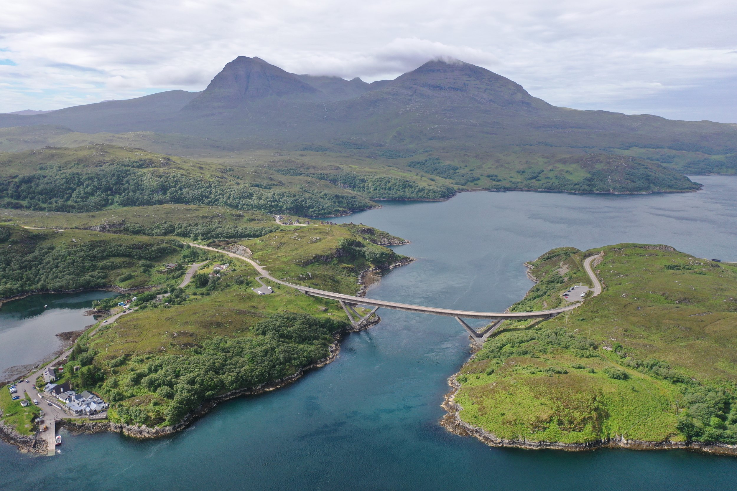 Aerial view of a bridge spanning a blue river with green hills and a mountain in the background, under a cloudy sky.