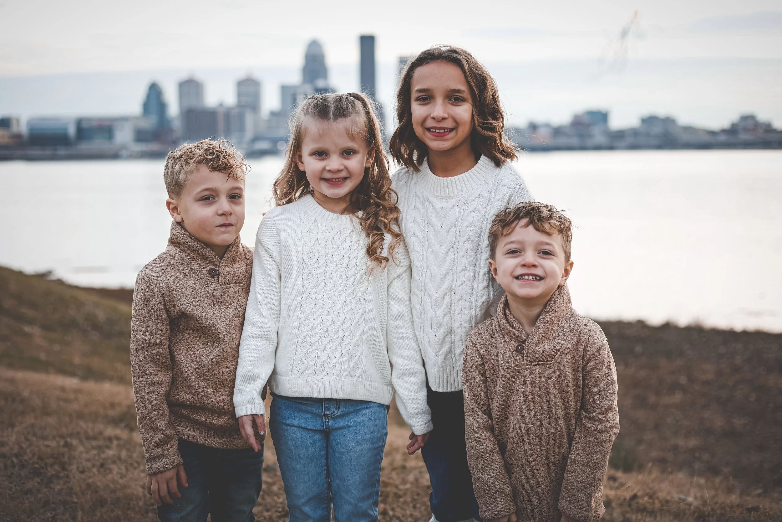 Four children posing together outdoors, wearing sweaters, with a city skyline in the background.