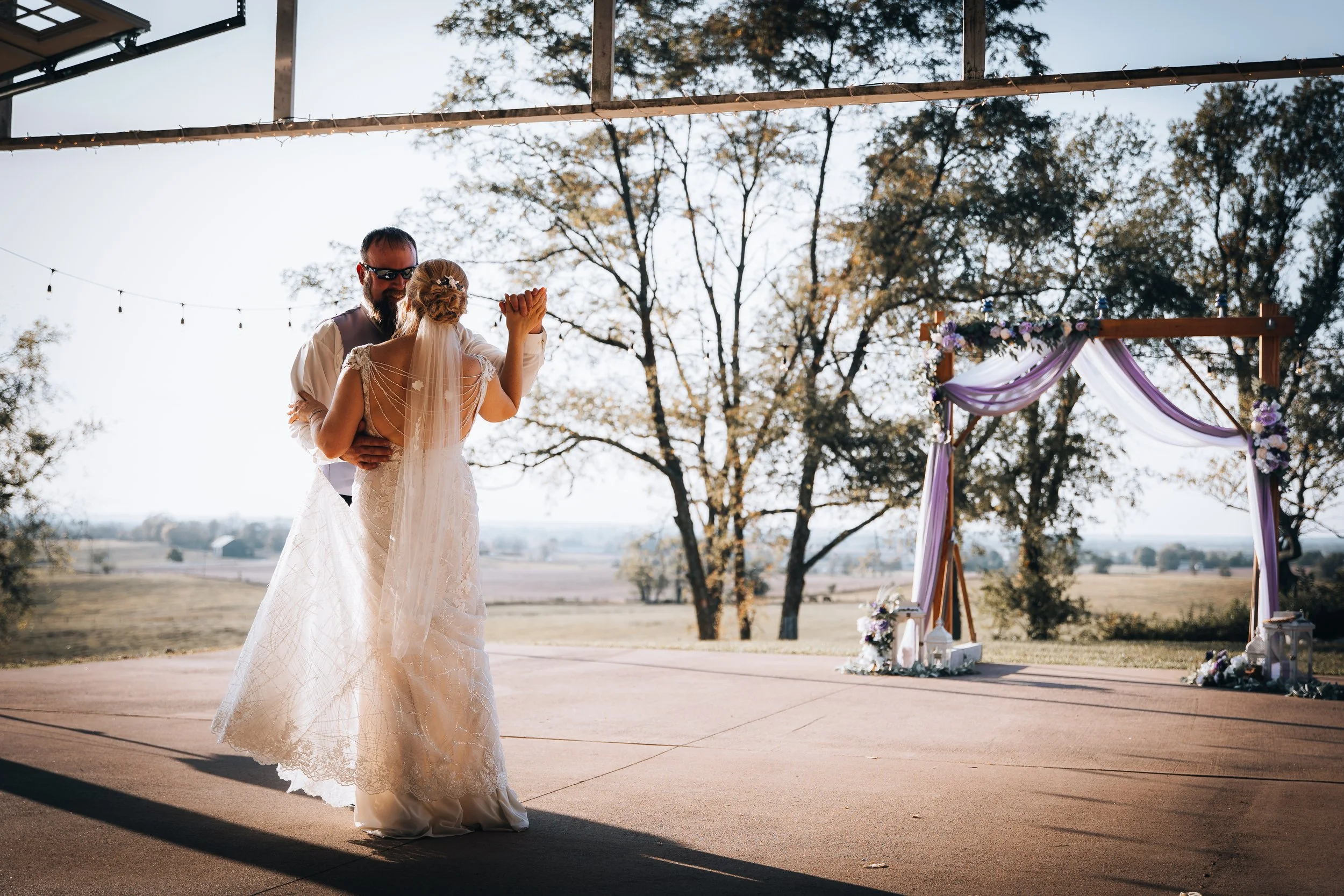 A couple dancing at an outdoor wedding ceremony under a decorated arch with purple fabric and flowers.