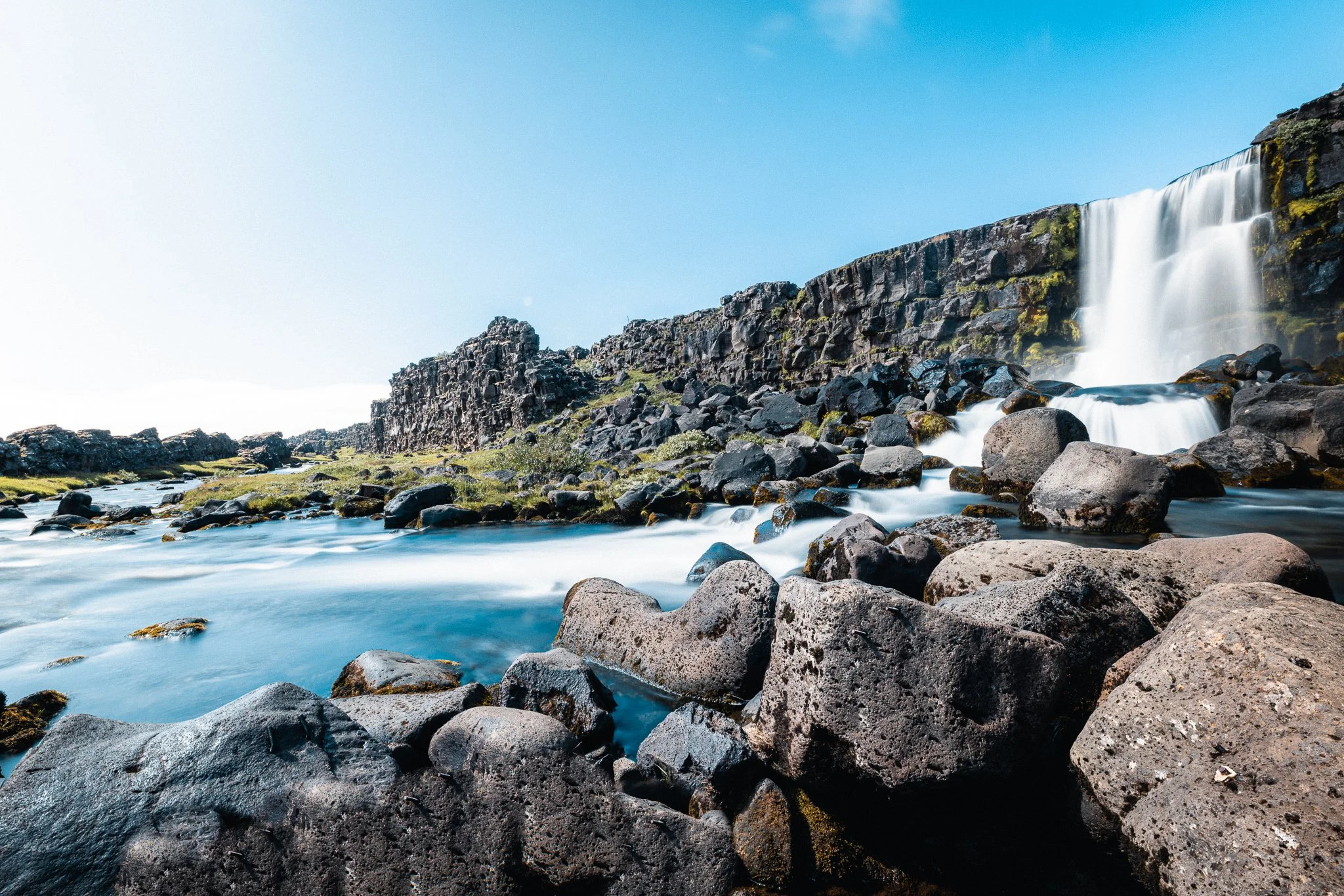 Waterfall cascading down rocky cliffs into a river, surrounded by moss-covered boulders and a clear blue sky.