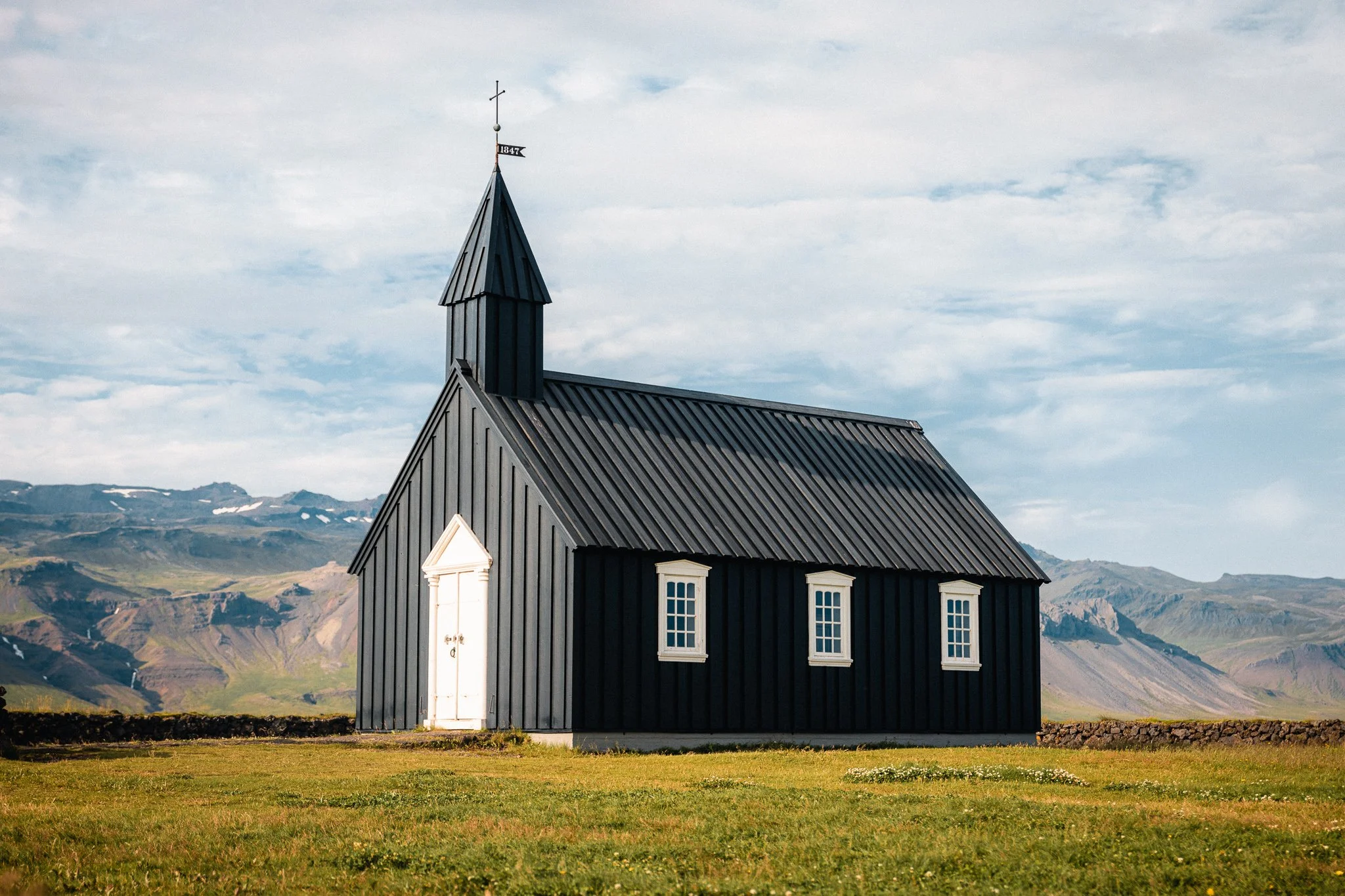 Black church with white doors and windows in a grassy field with mountain background and cloudy sky.