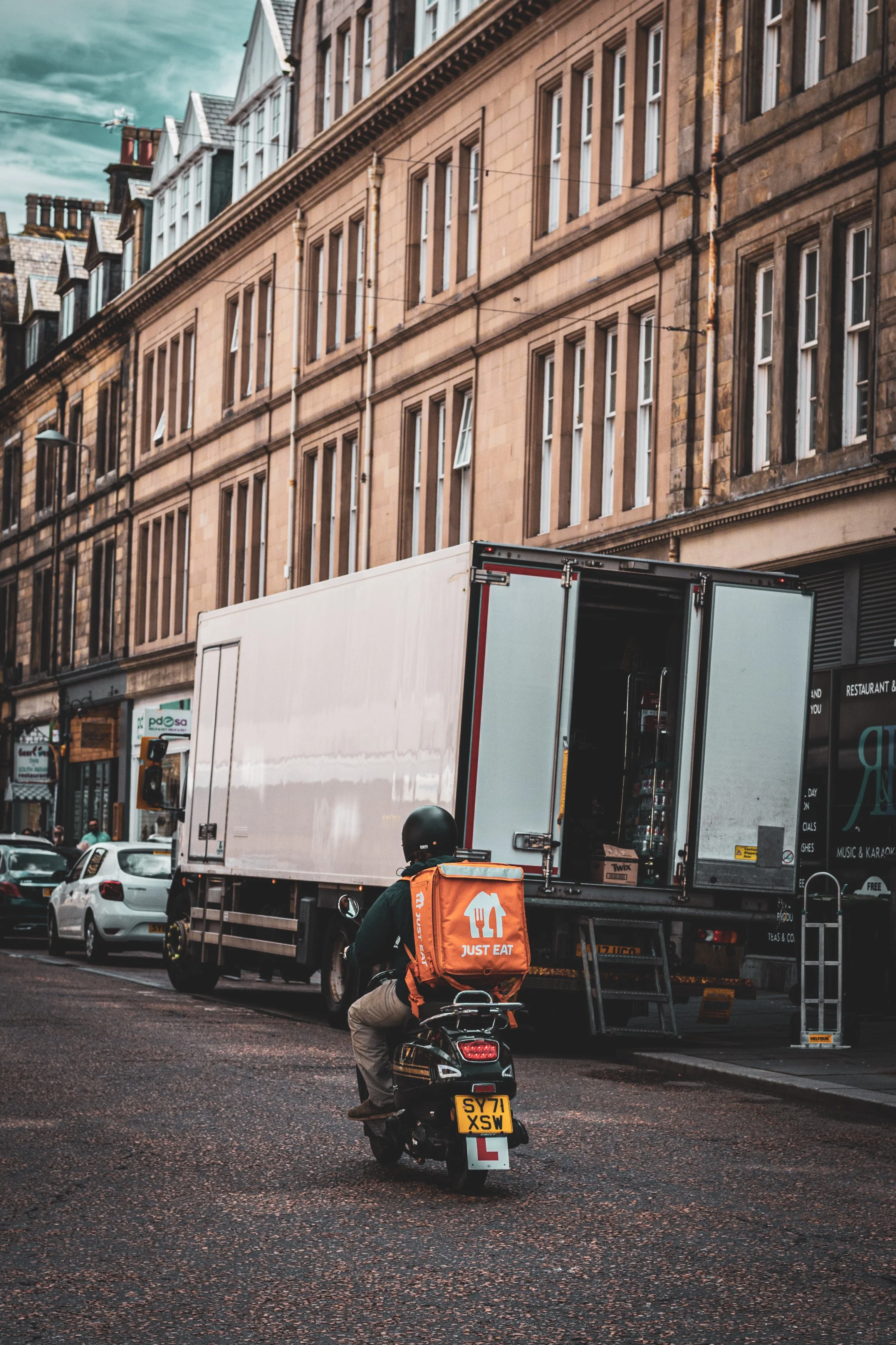 Delivery driver on a scooter with "Just Eat" backpack, passing a large truck on a city street.