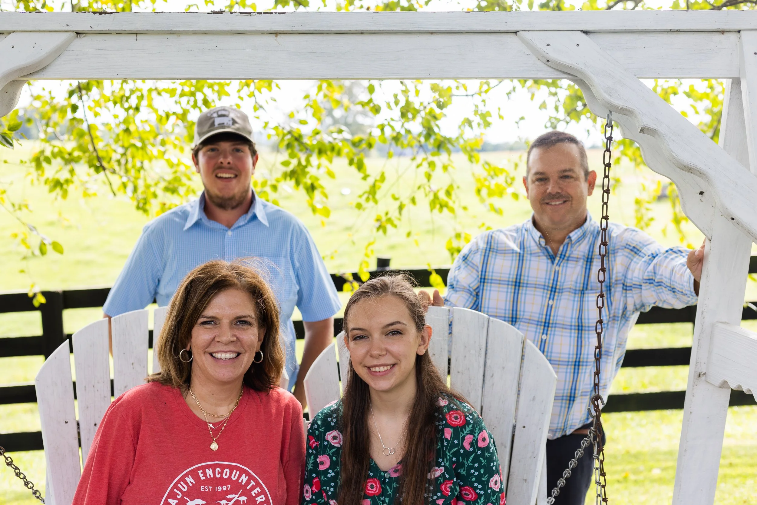 Four people smiling in a garden setting with two sitting on a white swing and two standing behind it. The background features leafy trees and a wooden fence.
