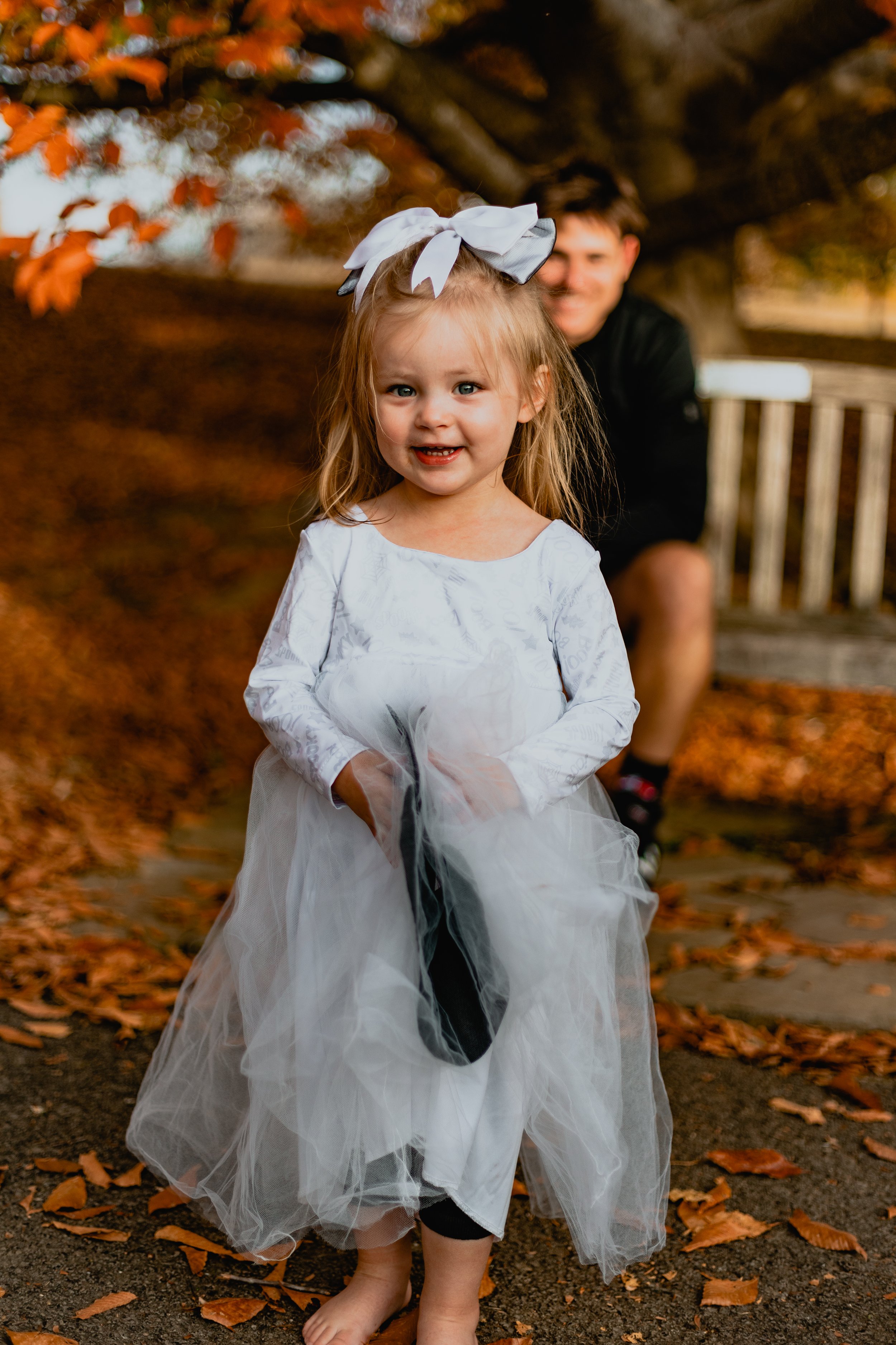 Young girl in a white dress with a large bow in her hair, standing on a path covered with autumn leaves, with a person smiling in the background.