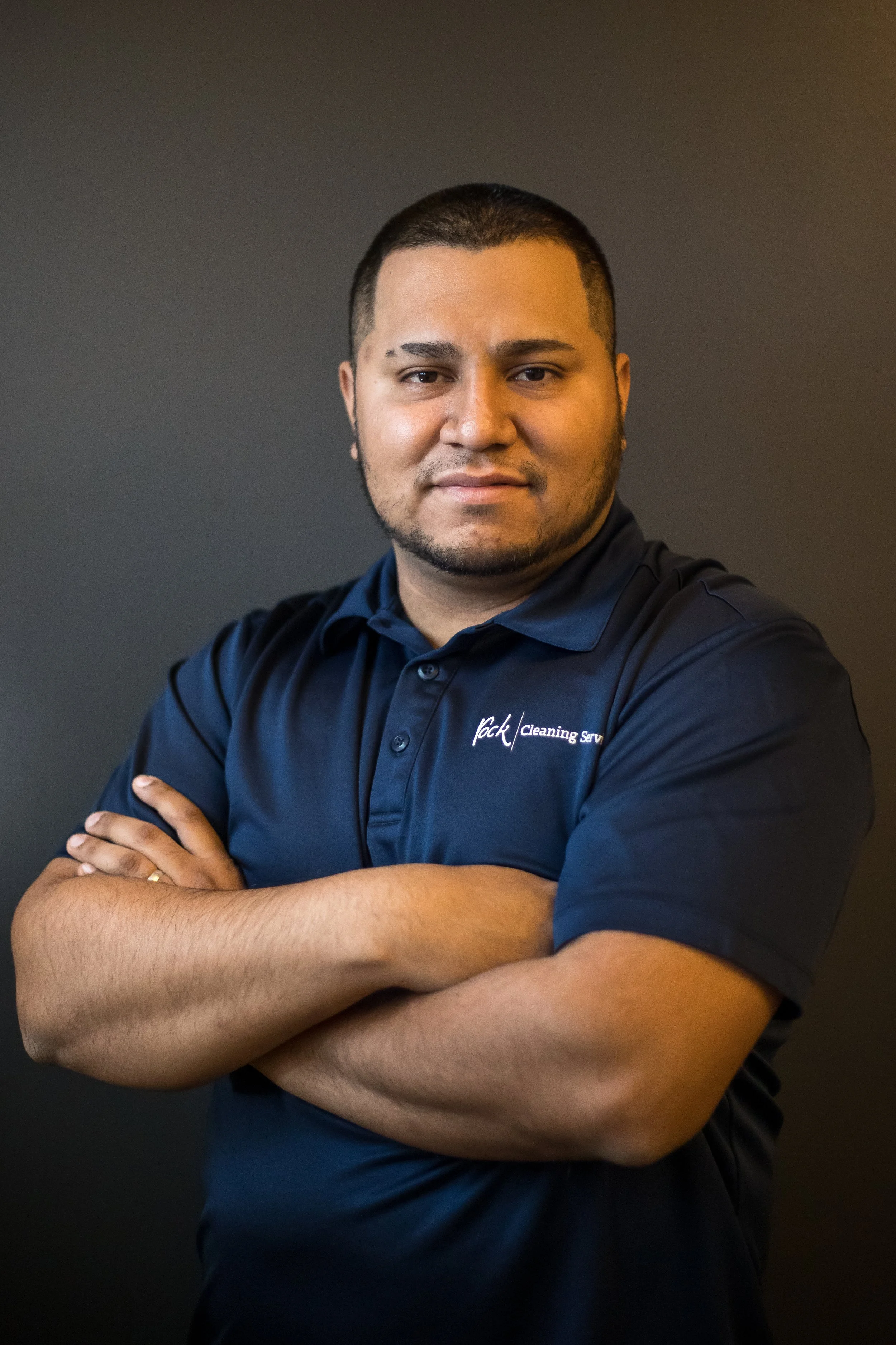 Man with beard crossing his arms and wearing a navy polo shirt against a dark background.