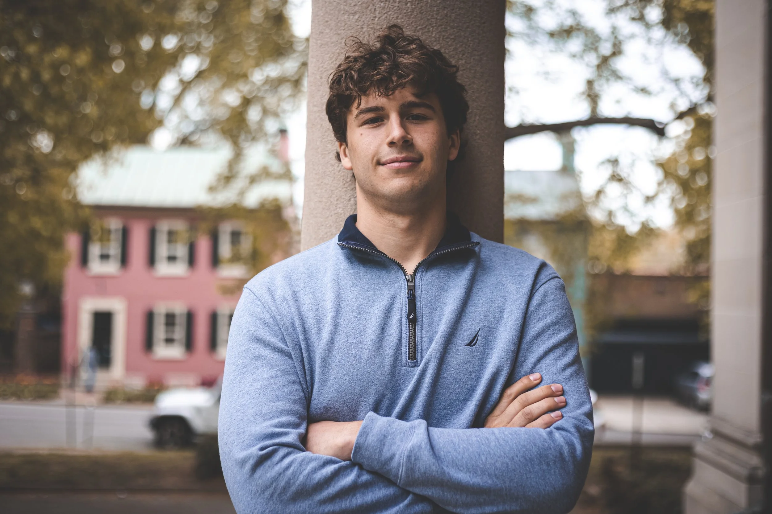 Man in blue sweater leaning against a column with arms crossed, in front of a blurred background of trees and a building.