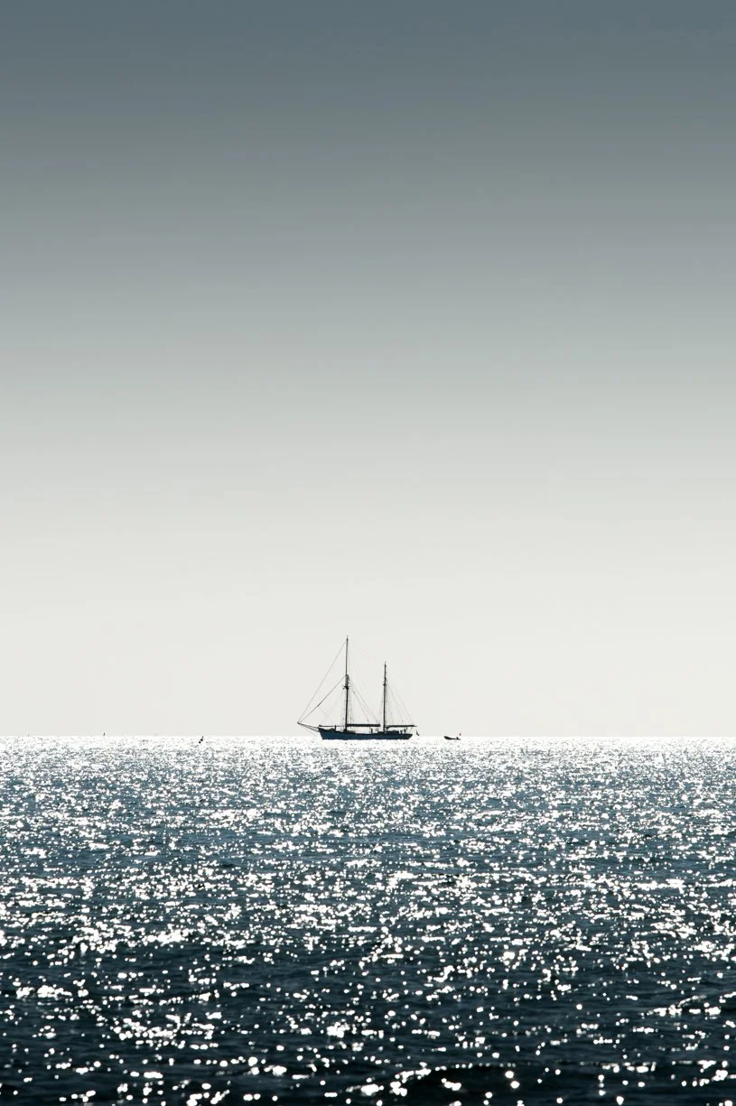 Un voilier naviguant sur une mer calme, avec un ciel clair et un soleil brillant qui reflète sur l'eau.