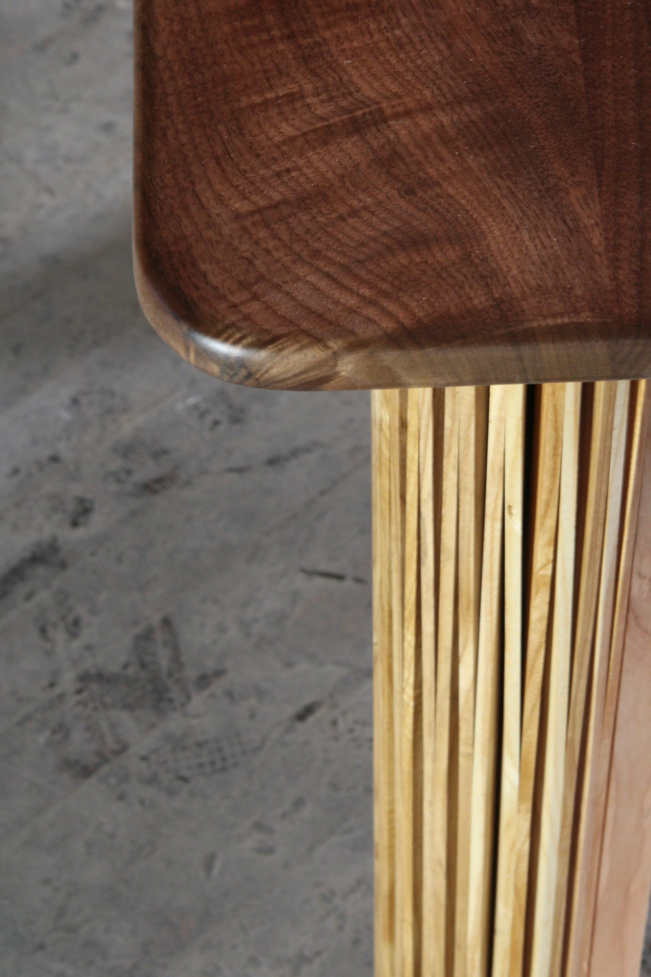 Close-up of a piece of wooden furniture, showing the corner of a round tabletop and vertical slats on the side, placed on a concrete floor.