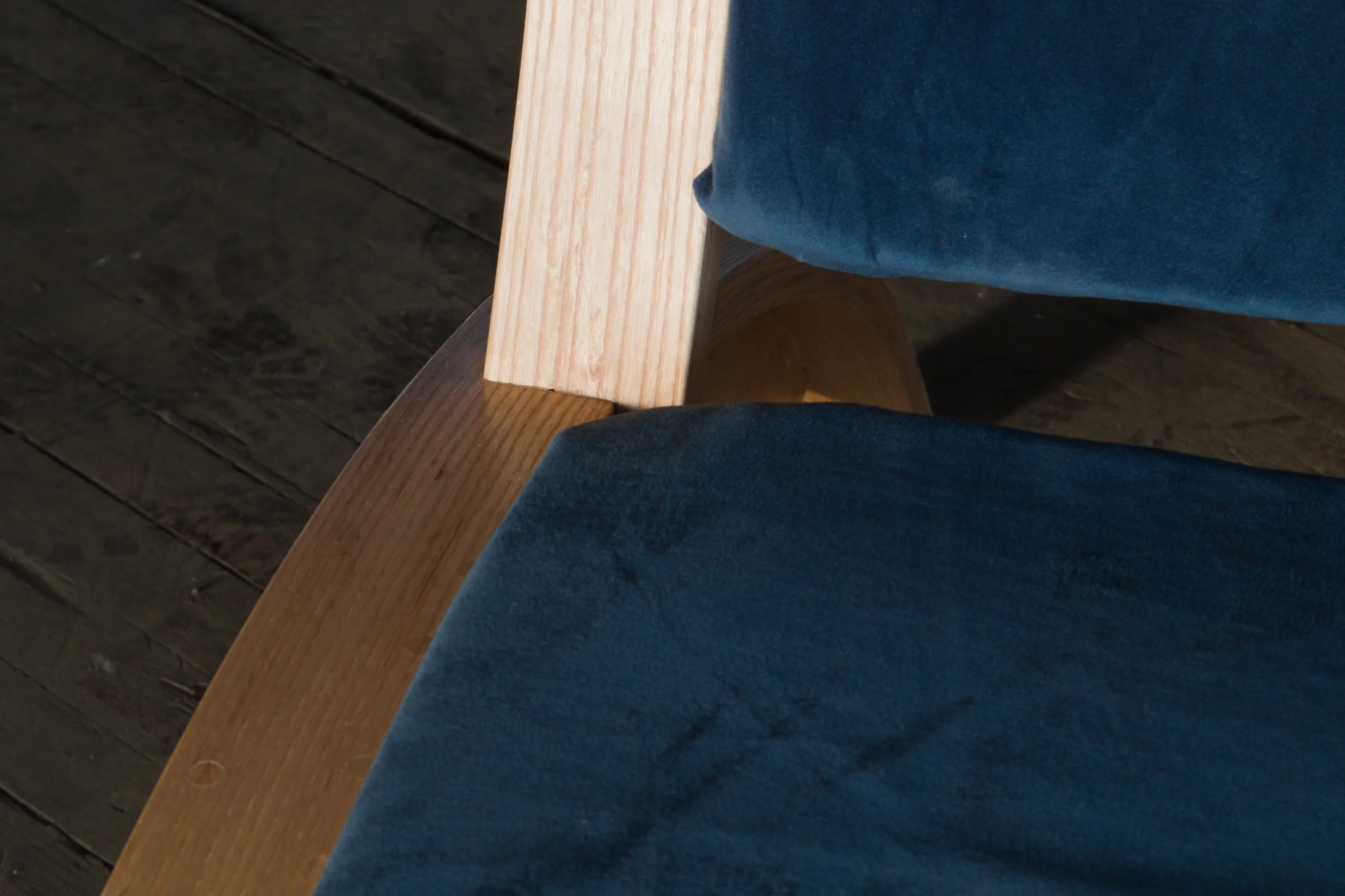 Close-up of a wooden chair with dark blue fabric cushion, showing the armrest and backrest, placed on a dark wooden floor.
