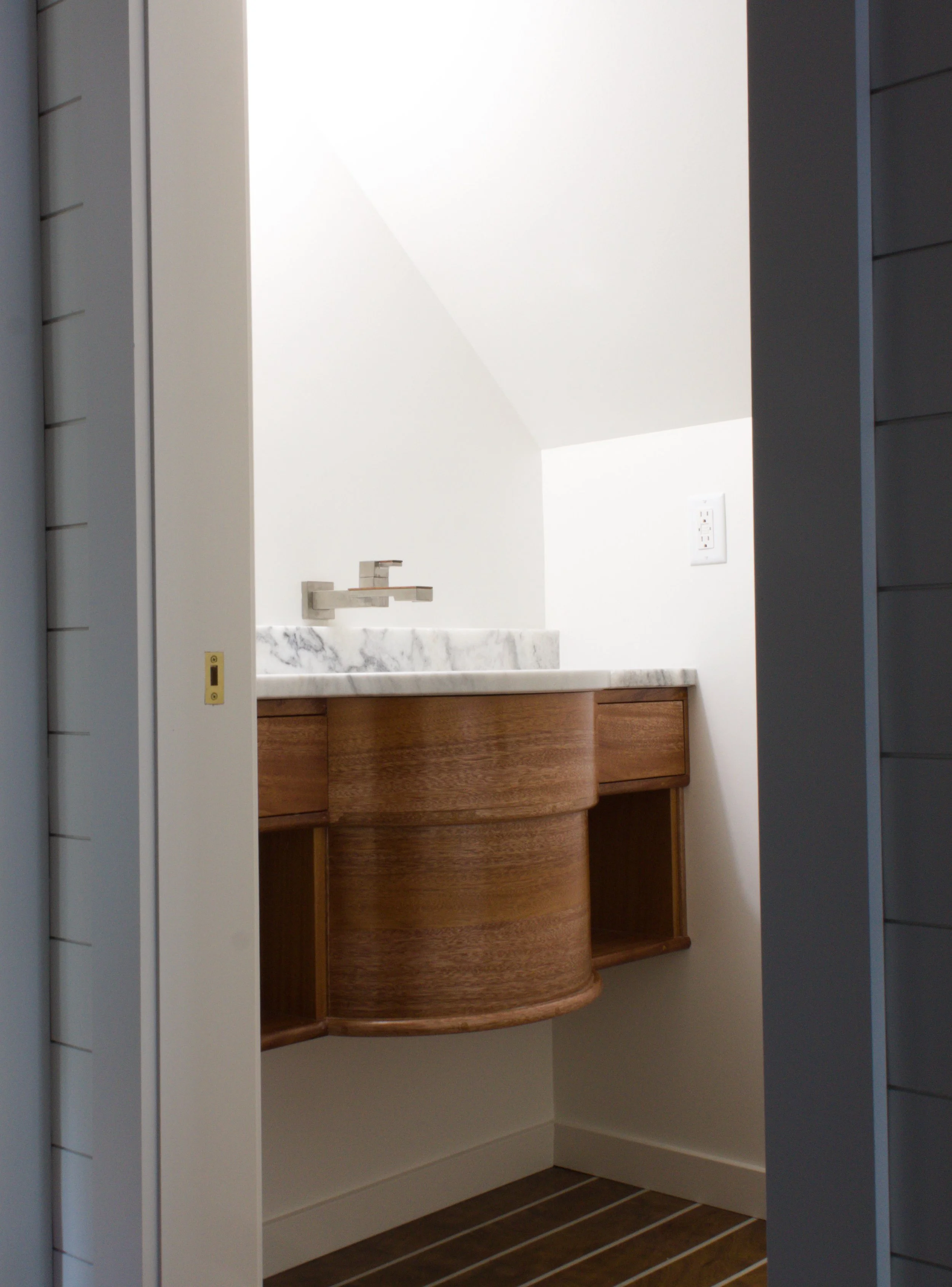 Small bathroom vanity with a marble countertop, modern faucet, and wooden cabinet, seen through an open door.
