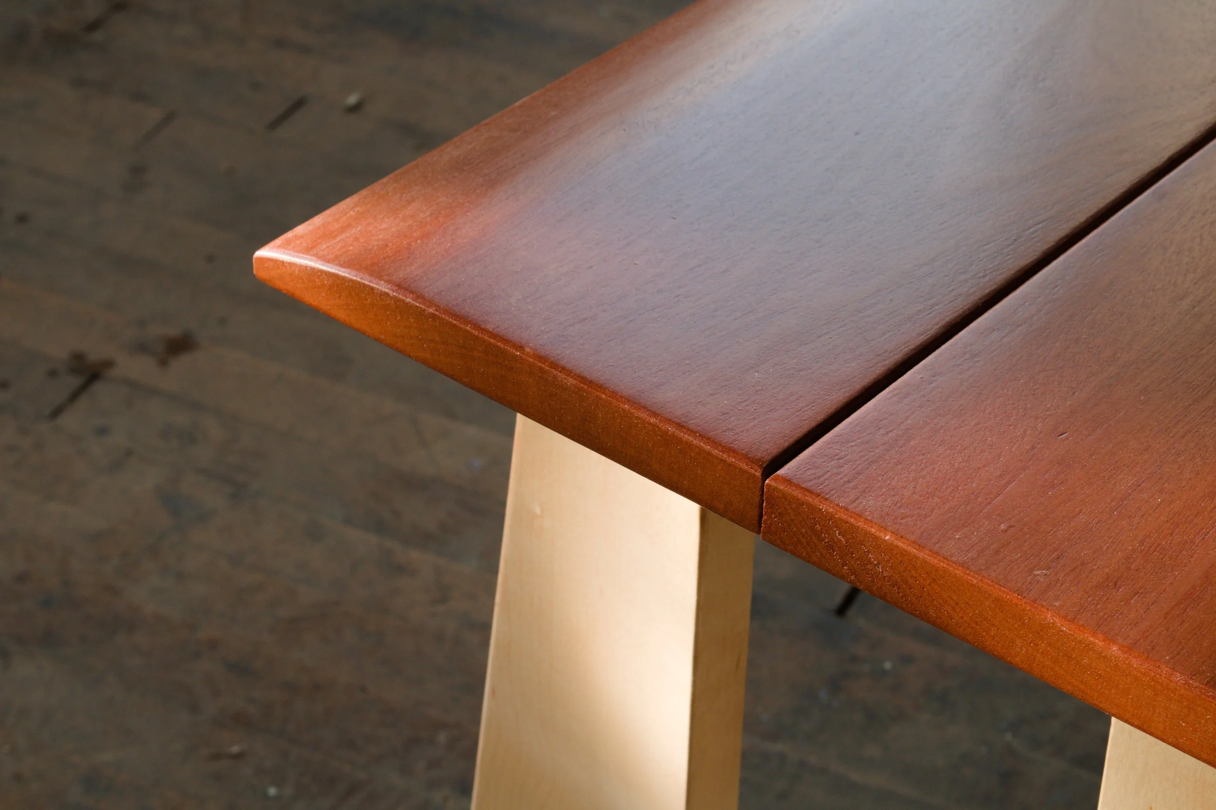 Close-up of a wooden table with a reddish-brown polished surface and light-colored legs, on a dark wooden floor.