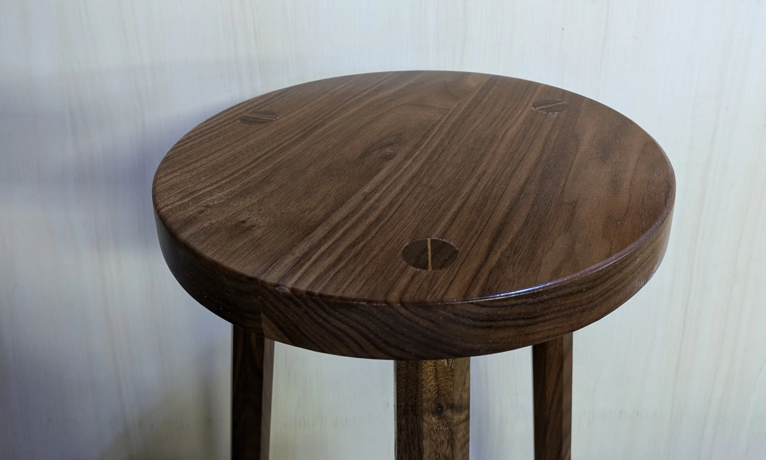 Close-up of a round wooden side table with a smooth surface and visible wood grain, against a plain light-colored wall.