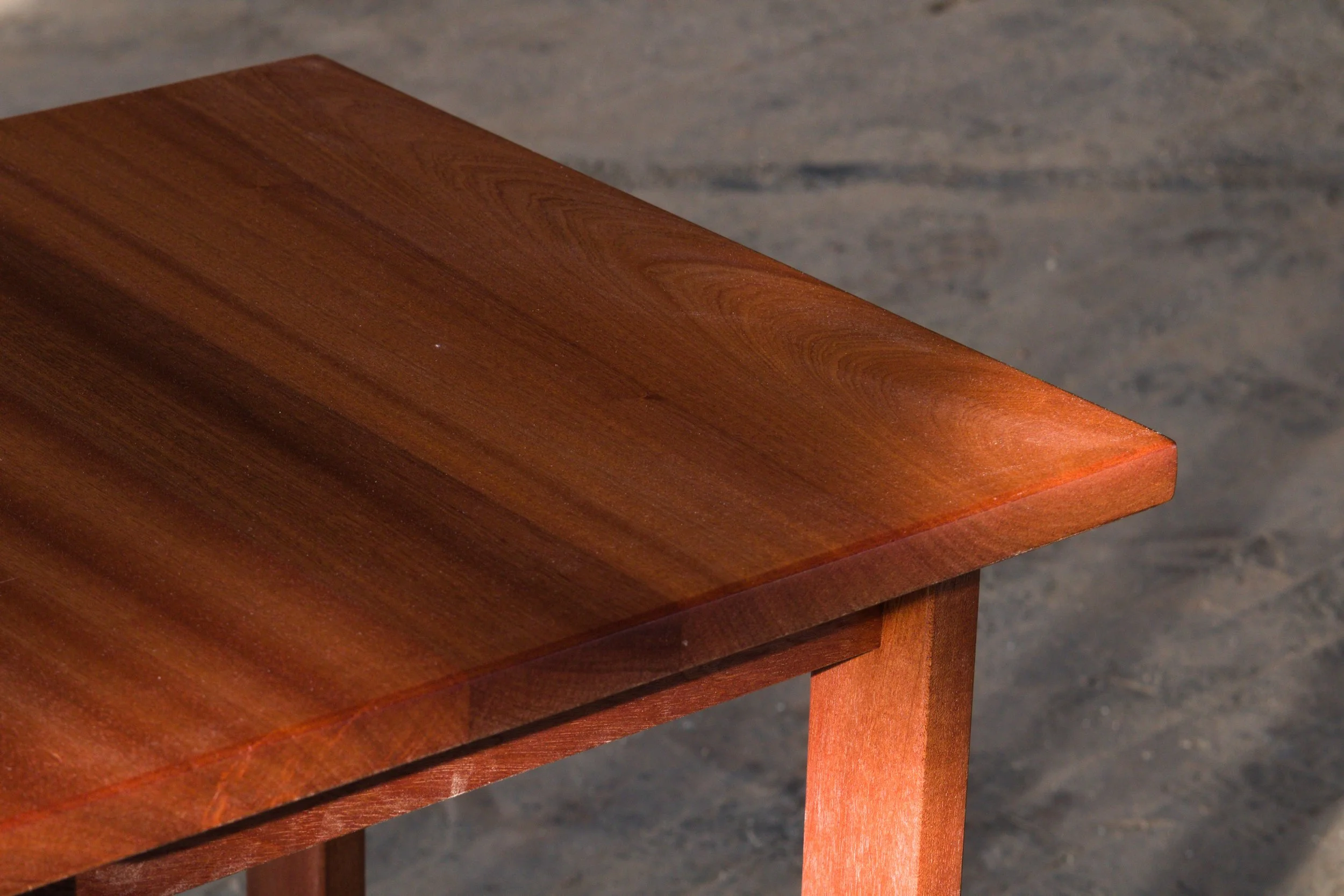 Close-up of a wooden table with a polished, reddish-brown finish, showing the edge and part of the leg on a gray concrete floor.