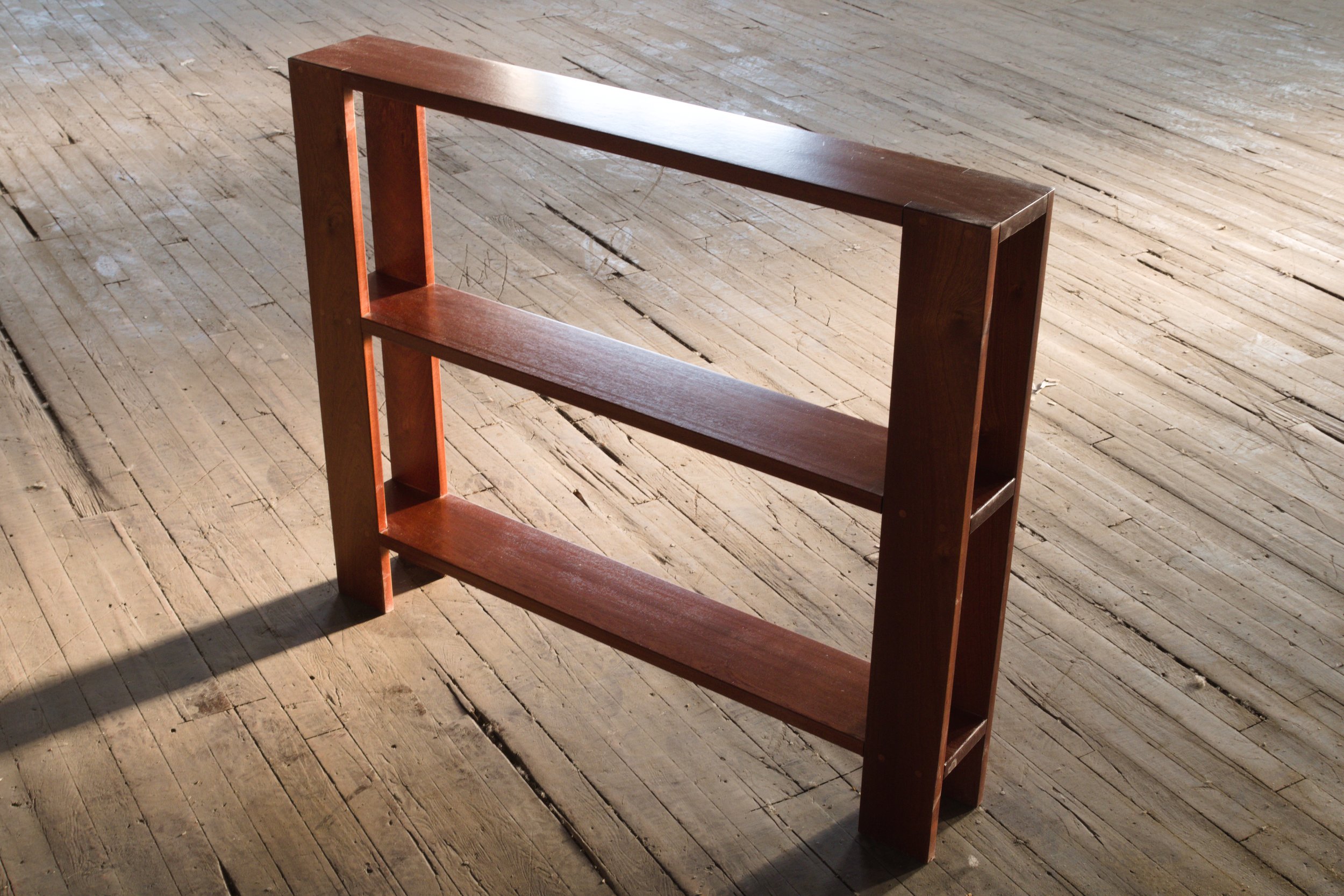 A three-tier wooden bookshelf with a reddish-brown finish on a wooden floor.