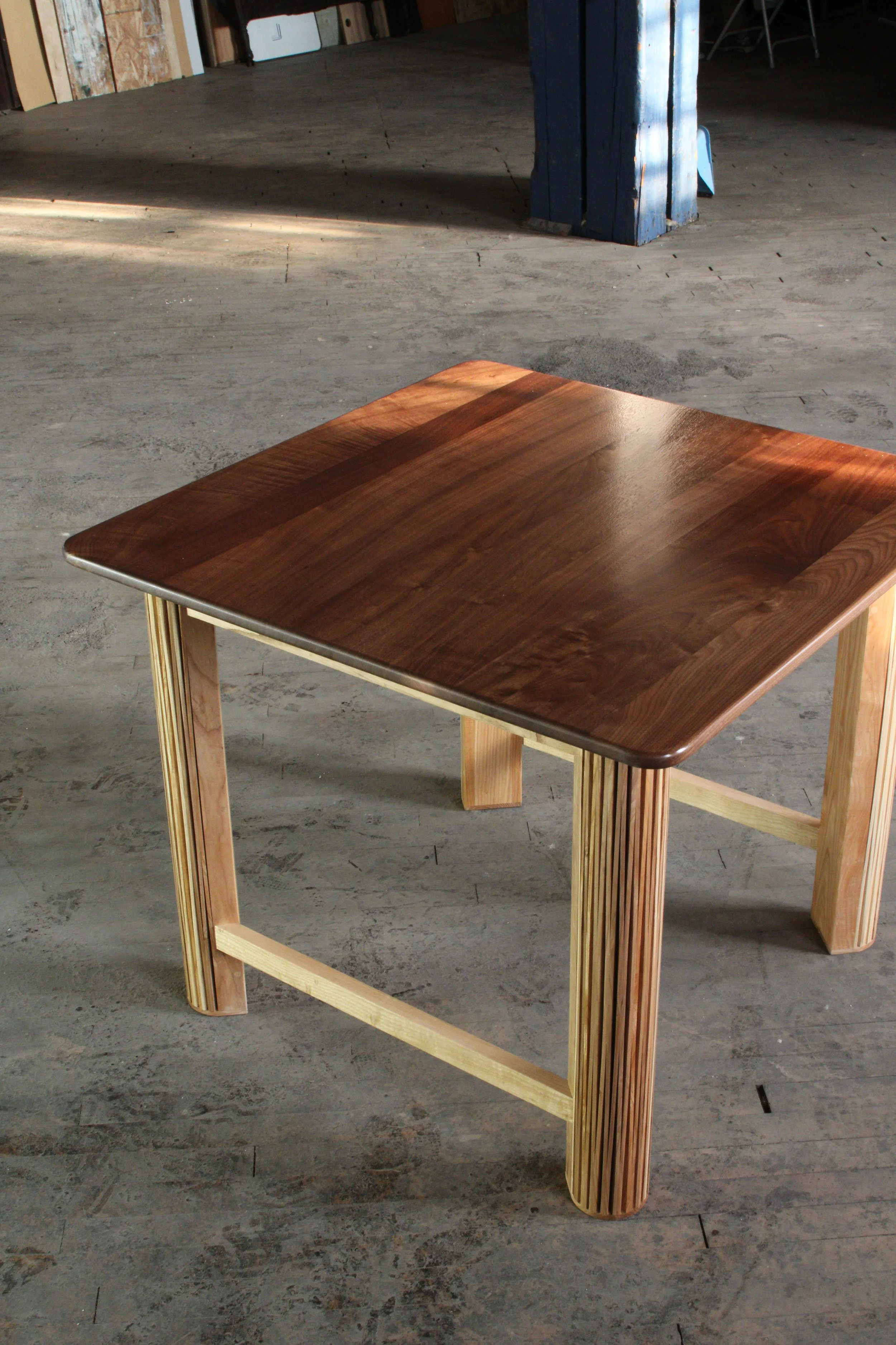 A square wooden table with a polished dark top and light wood legs, situated on a concrete floor inside an unfinished room.
