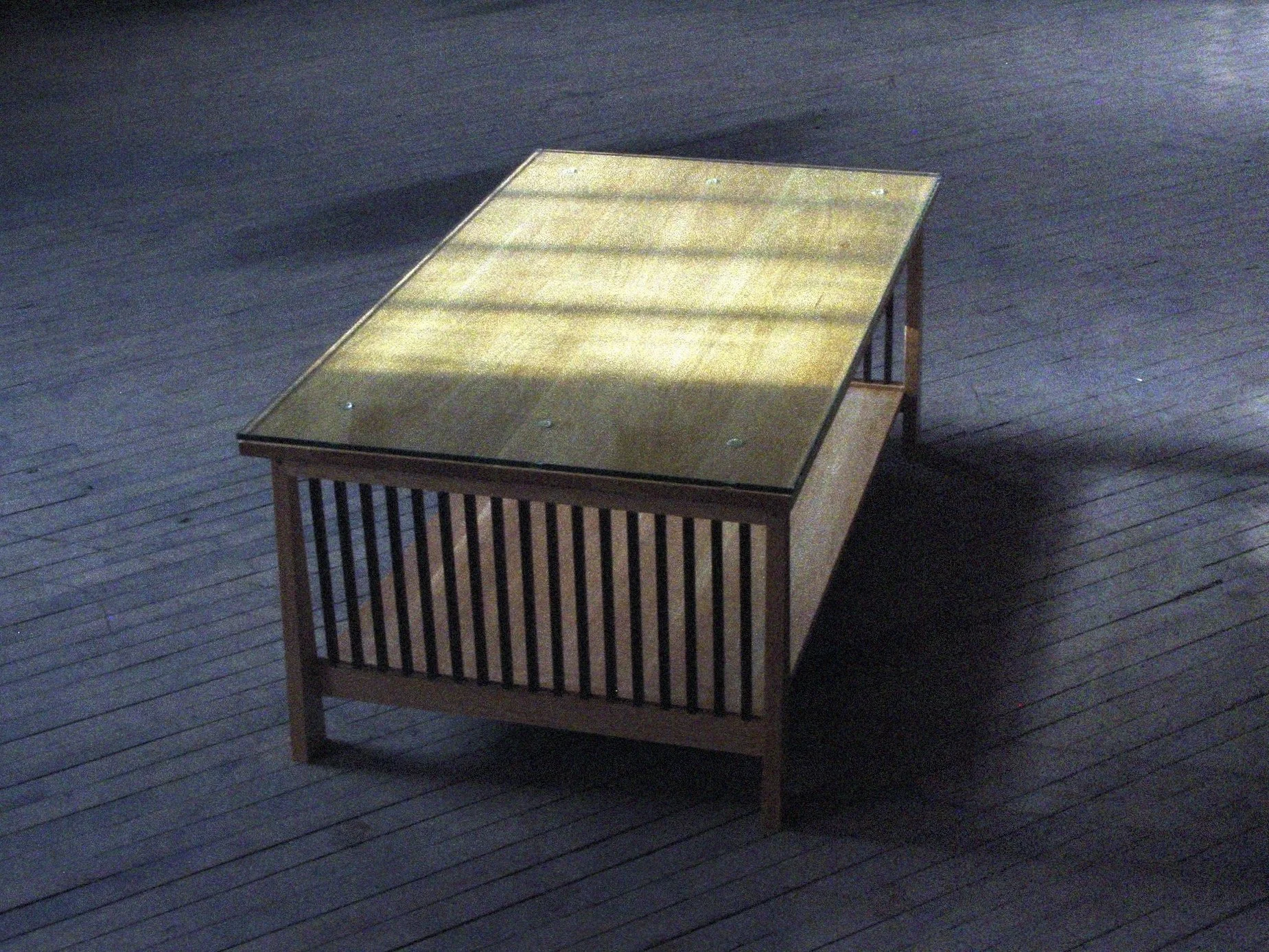 A wooden table with a glass top, placed on a dark wooden floor, with shadows cast on it.