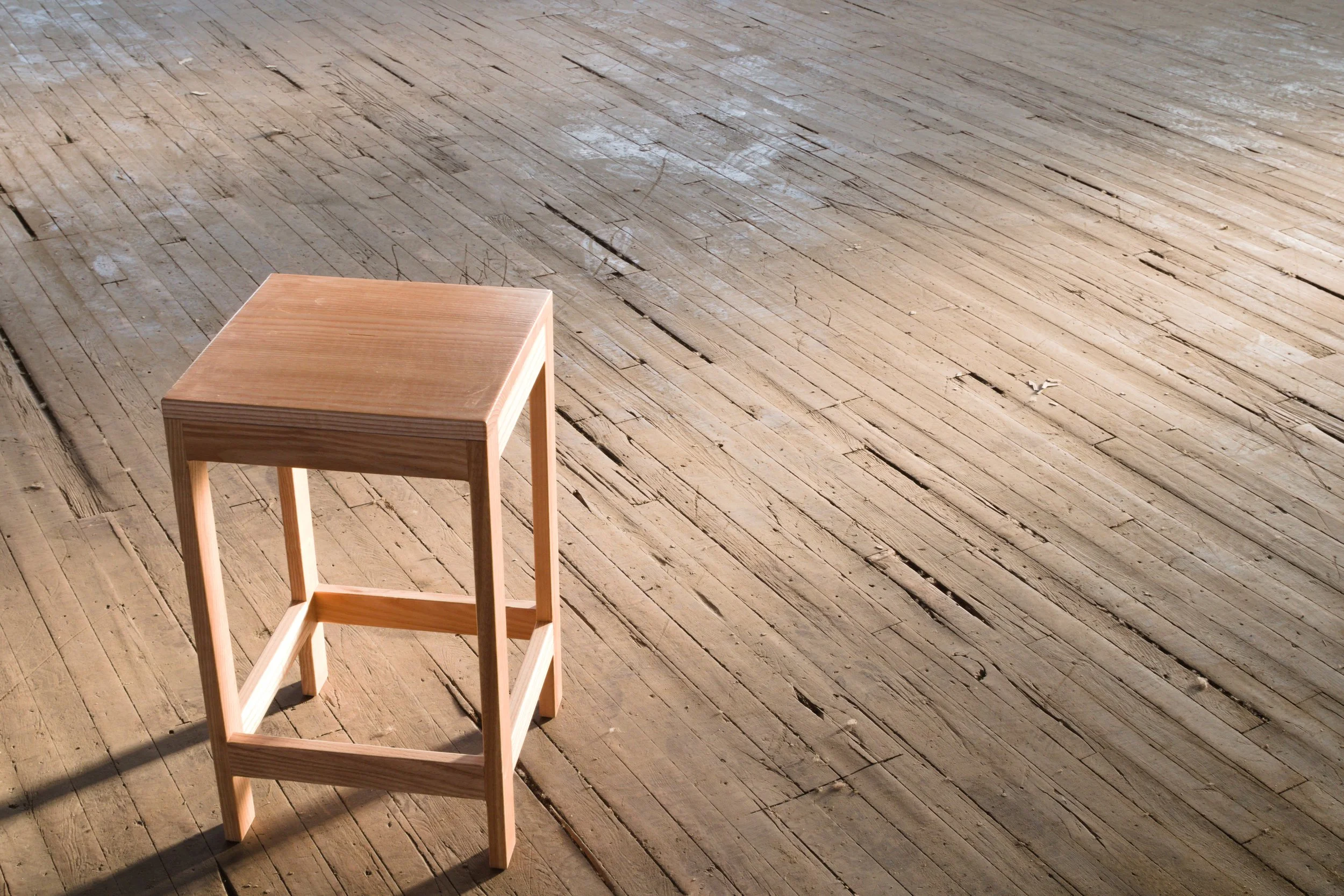 A simple wooden stool is placed on a rustic wooden floor with natural light casting shadows.