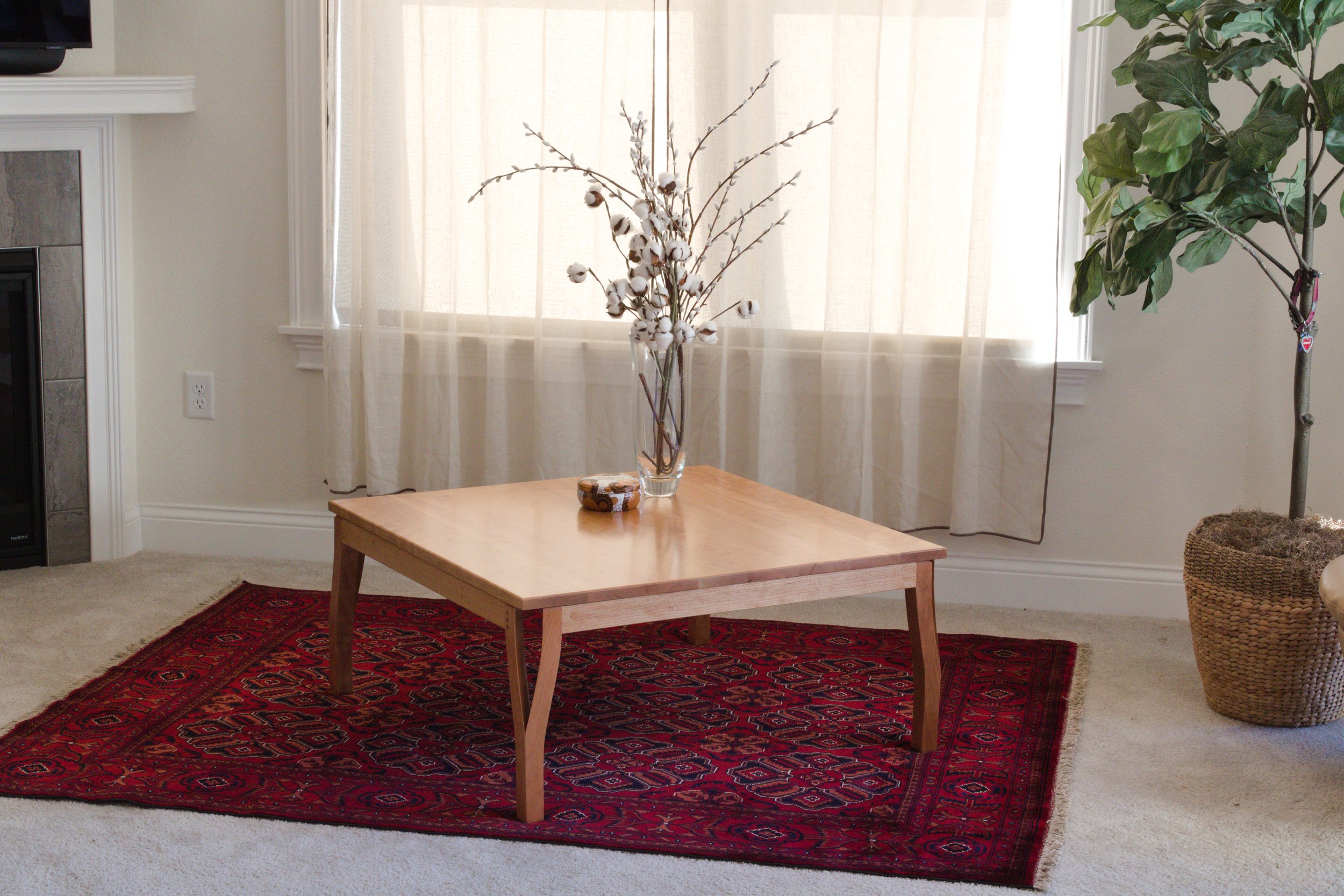 A wooden coffee table on a red patterned rug in a living room, with a glass vase of cotton branch and a small container on top, beige curtains, a potted plant, and part of a fireplace.