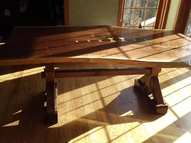 A wooden dining table with a polished dark finish, located near large windows letting in sunlight, with shadows cast on the floor.