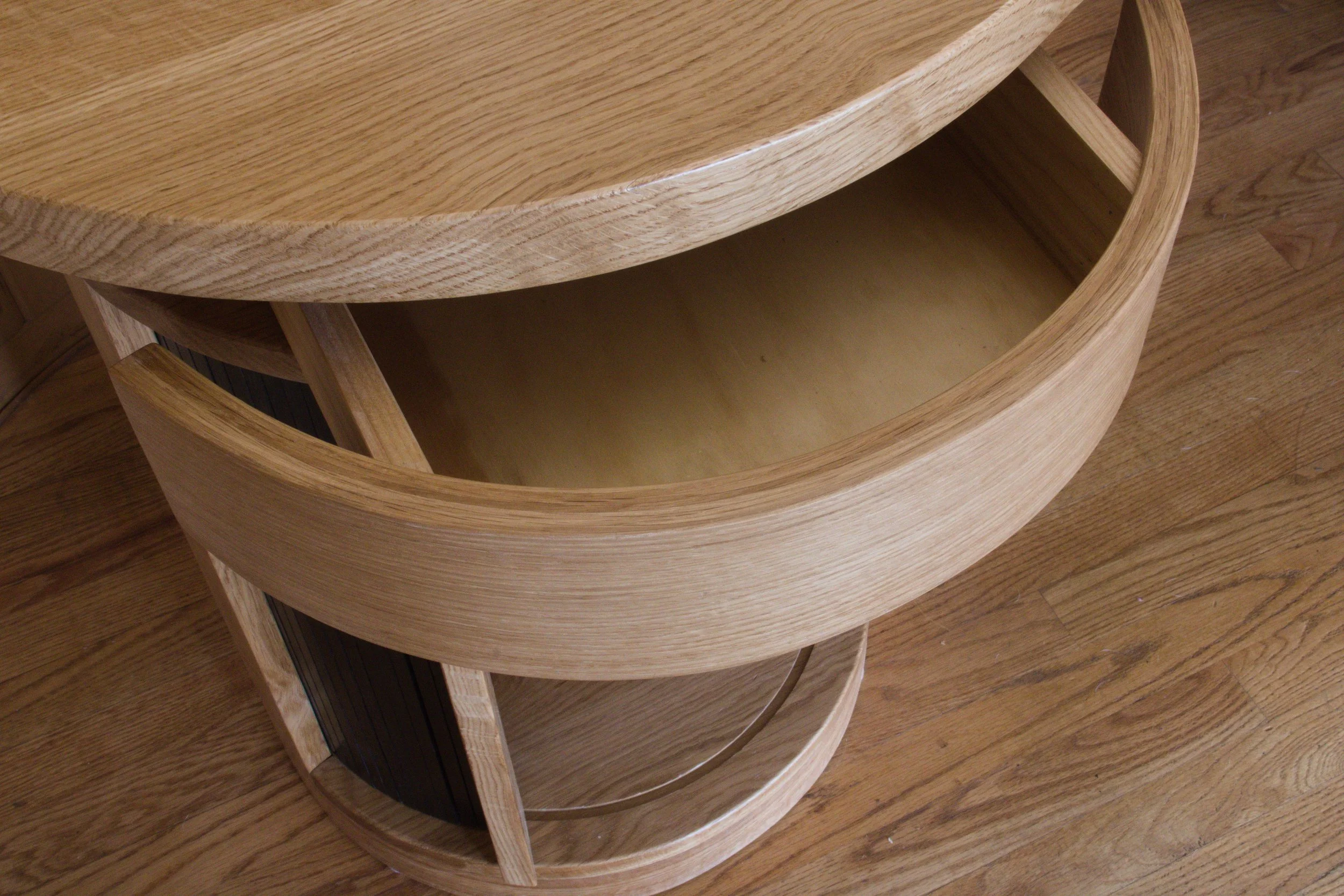 Wooden oval-shaped piece of furniture, possibly a cabinet or shelving unit, with a drawer pulled out, showing a light-colored interior and a curved wooden top, on a hardwood floor.