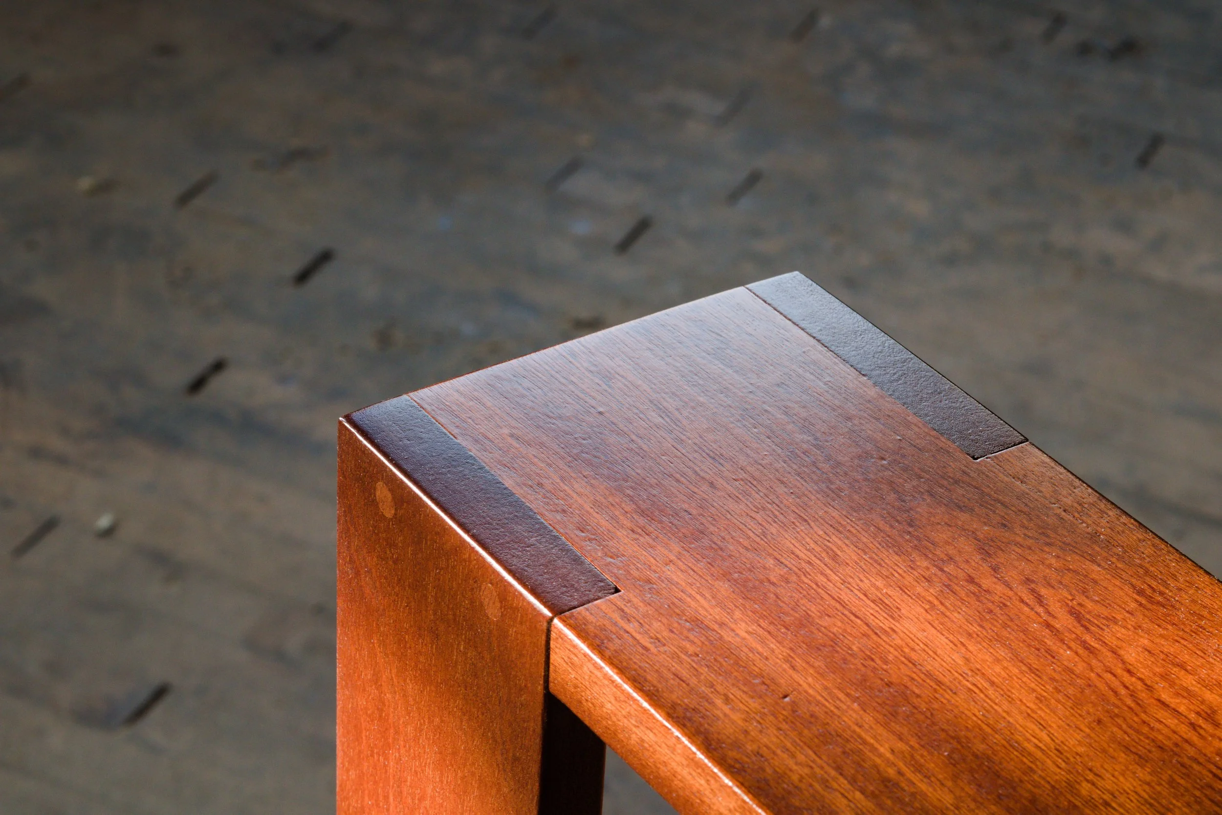 Close-up of the corner of a wooden furniture piece with a dark accent on the edge, set against a blurry dark wooden floor background.