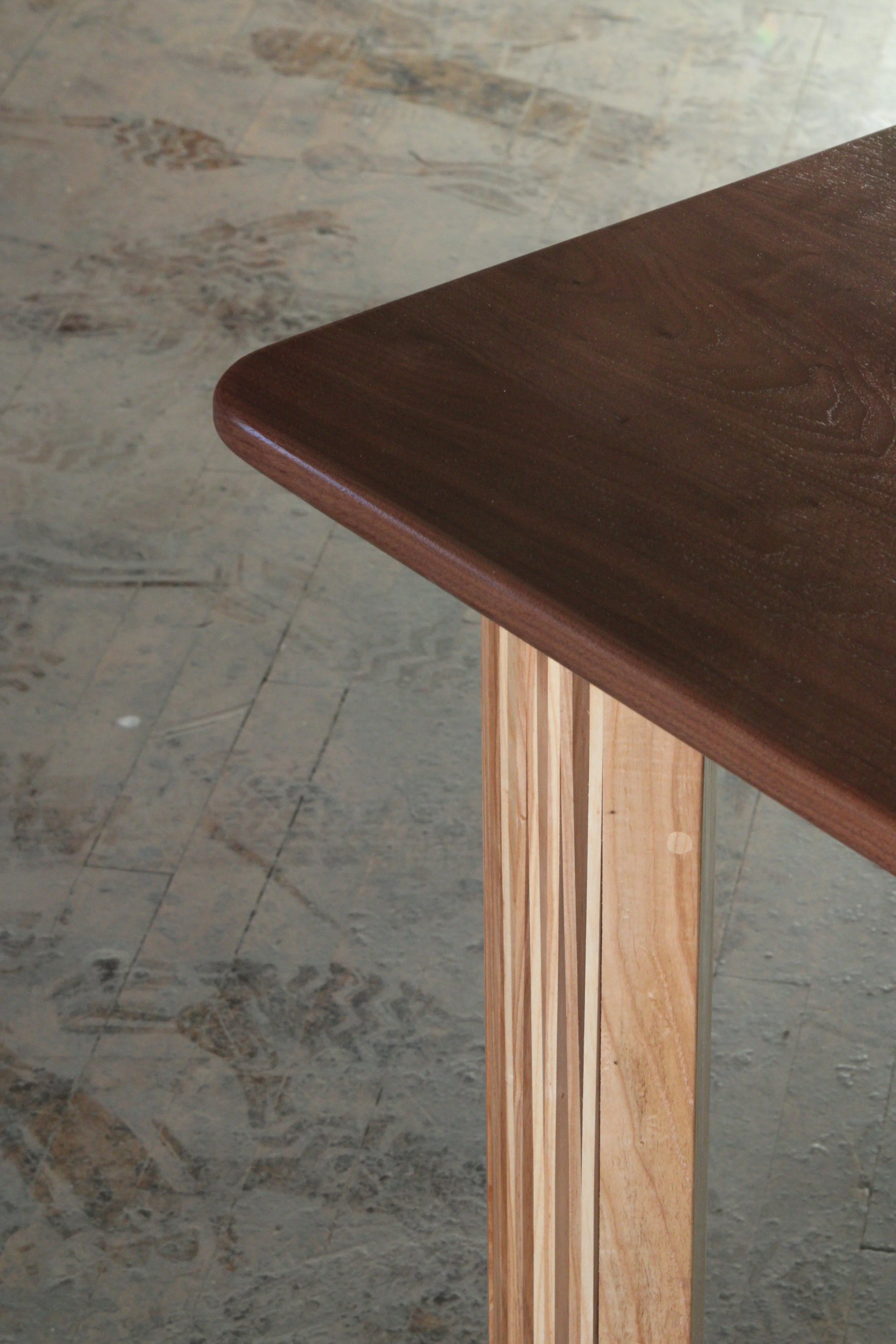 Close-up of a corner of a wooden table with a dark brown top and lighter wood support, on a textured stone floor.