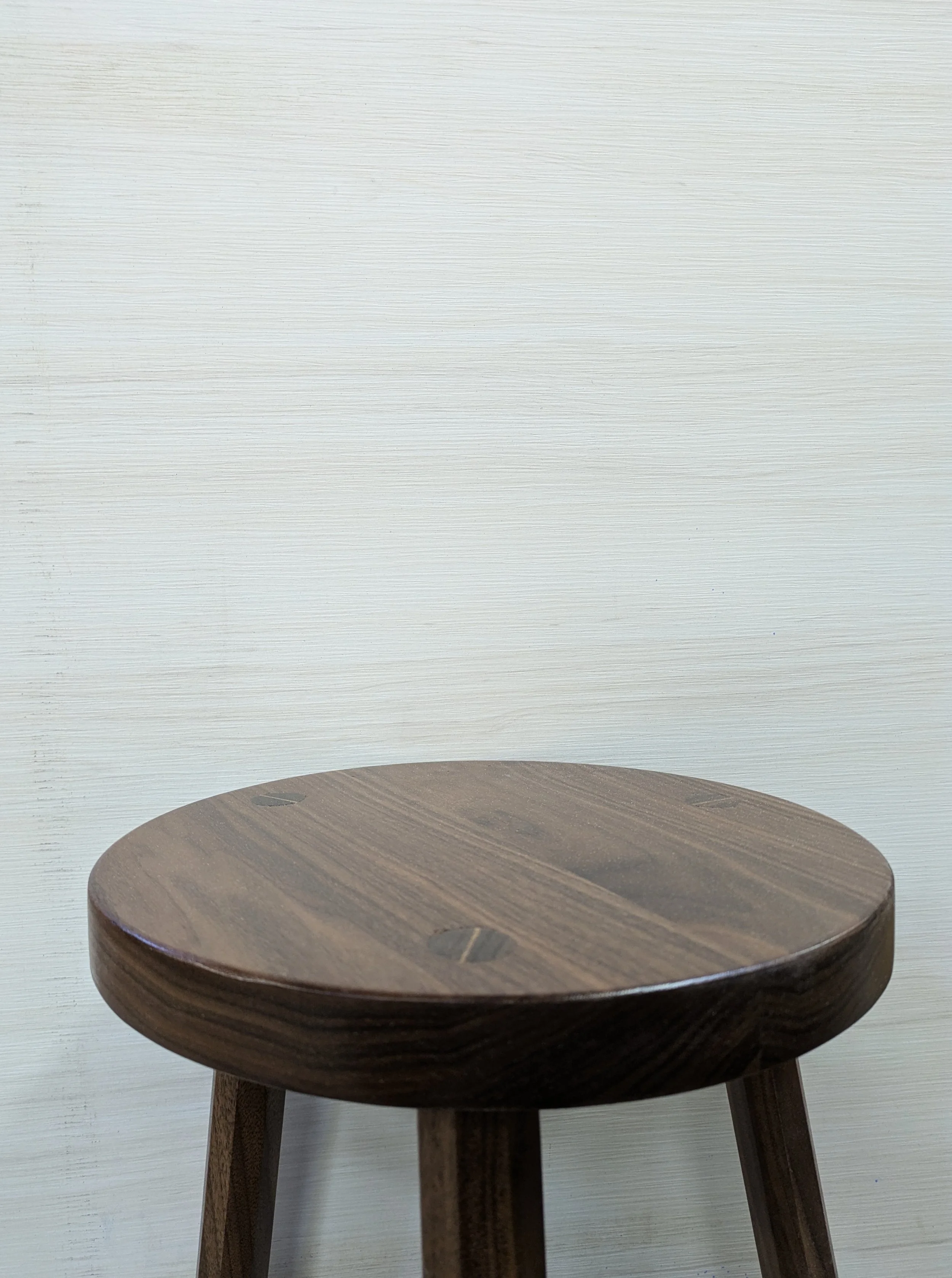 A wooden stool with three legs in front of a white wall.