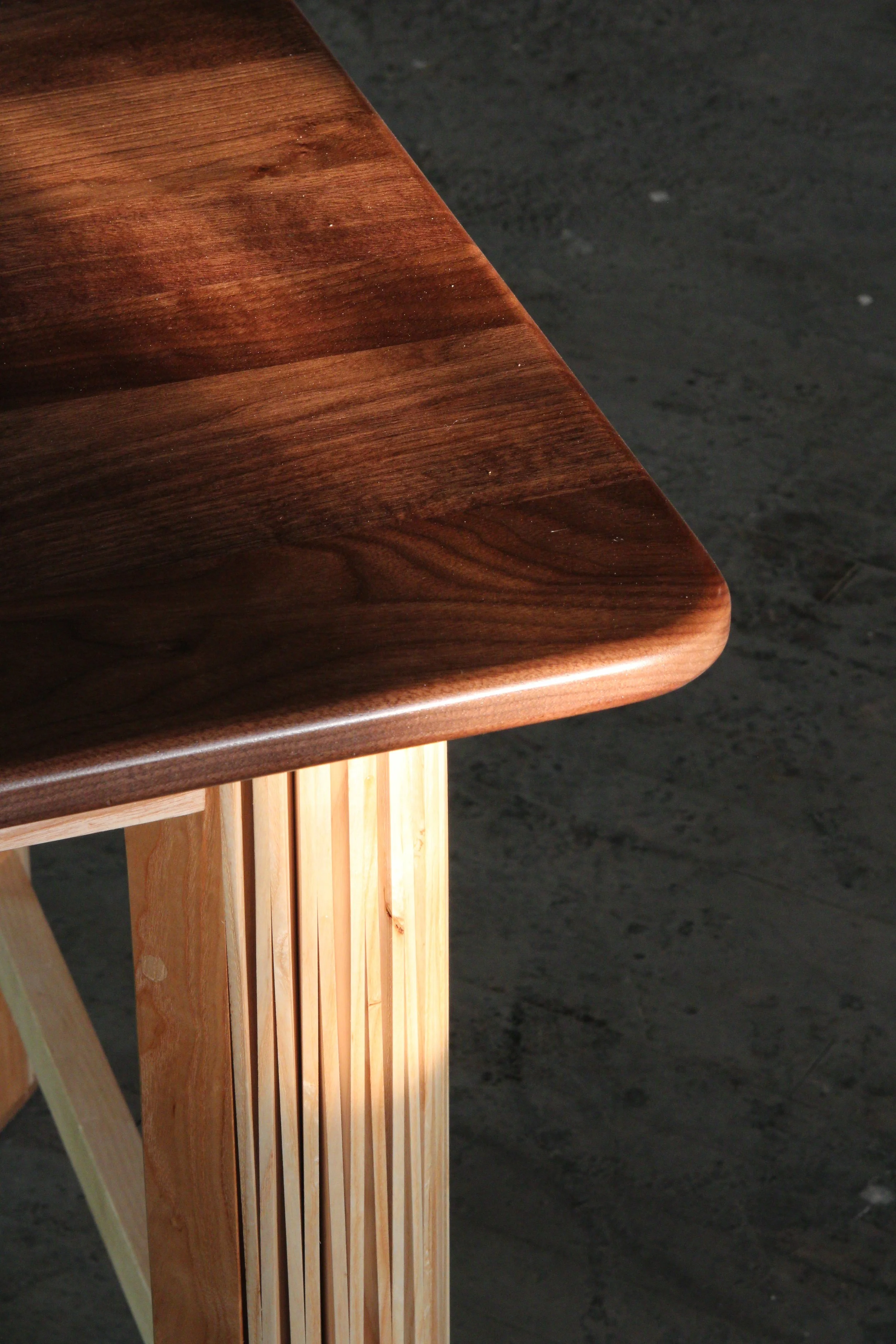 Close-up of a high-gloss polished wooden tabletop with rounded edges, supported by a light-colored wooden leg with evenly spaced vertical slats, set against a dark textured floor.