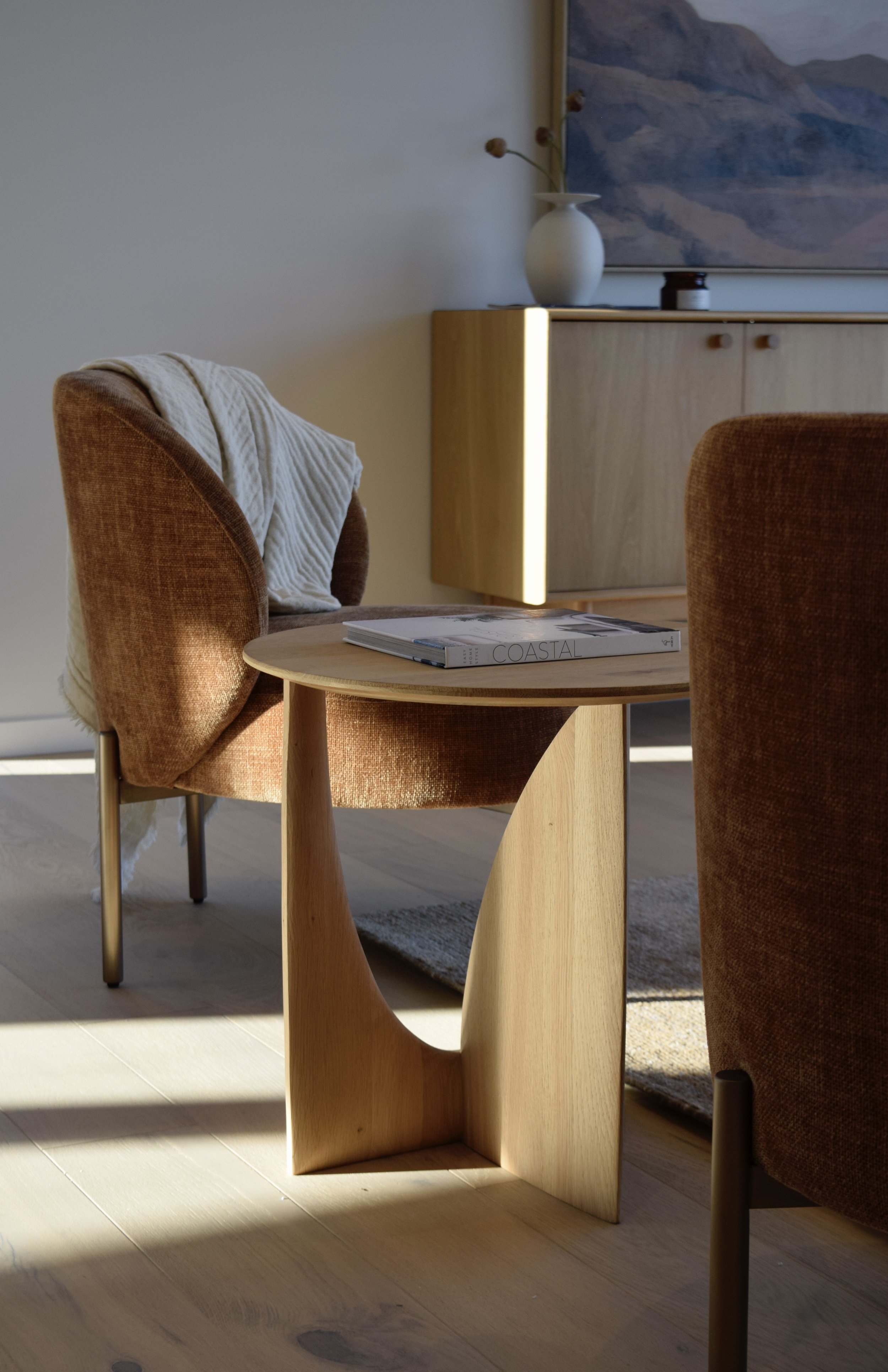 A cozy living room corner with two brown upholstered chairs, one with a white knit blanket draped over it. Between the chairs is a small round wooden side table with a magazine titled 'COASTAL.' In the background, a wooden cabinet has notes, a large 