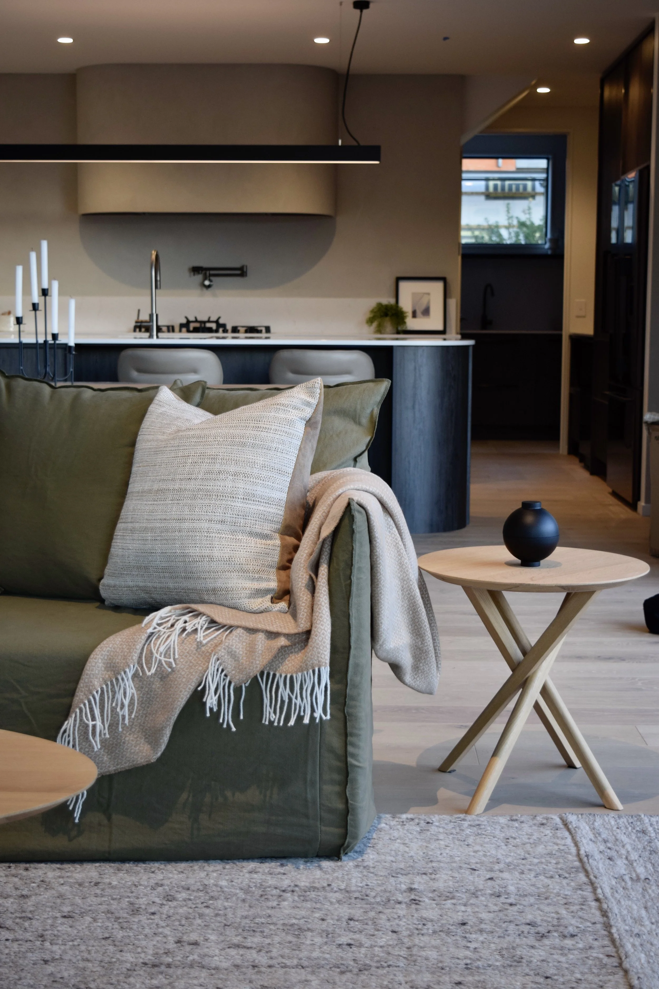 A living room with a sofa, pillows, a small wooden side table, and a cozy blanket. In the background, a modern kitchen with bar stools, a dark kitchen island, and a window with a view outside.