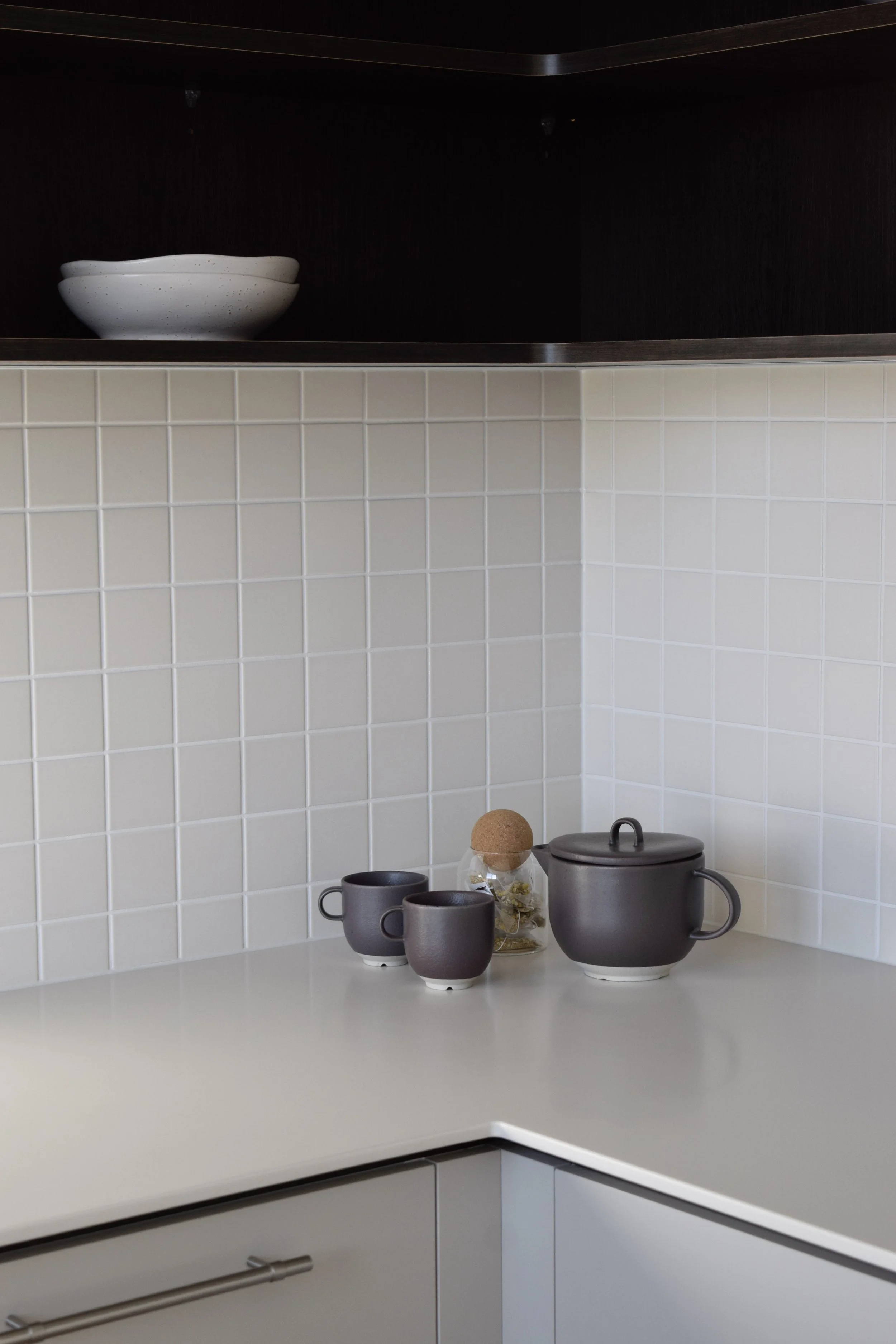 Kitchen corner with white tiled wall, white countertop, dark upper shelf with an empty white bowl, and a collection of gray ceramic mugs, teapot, and a small jar with a cork lid on the counter.