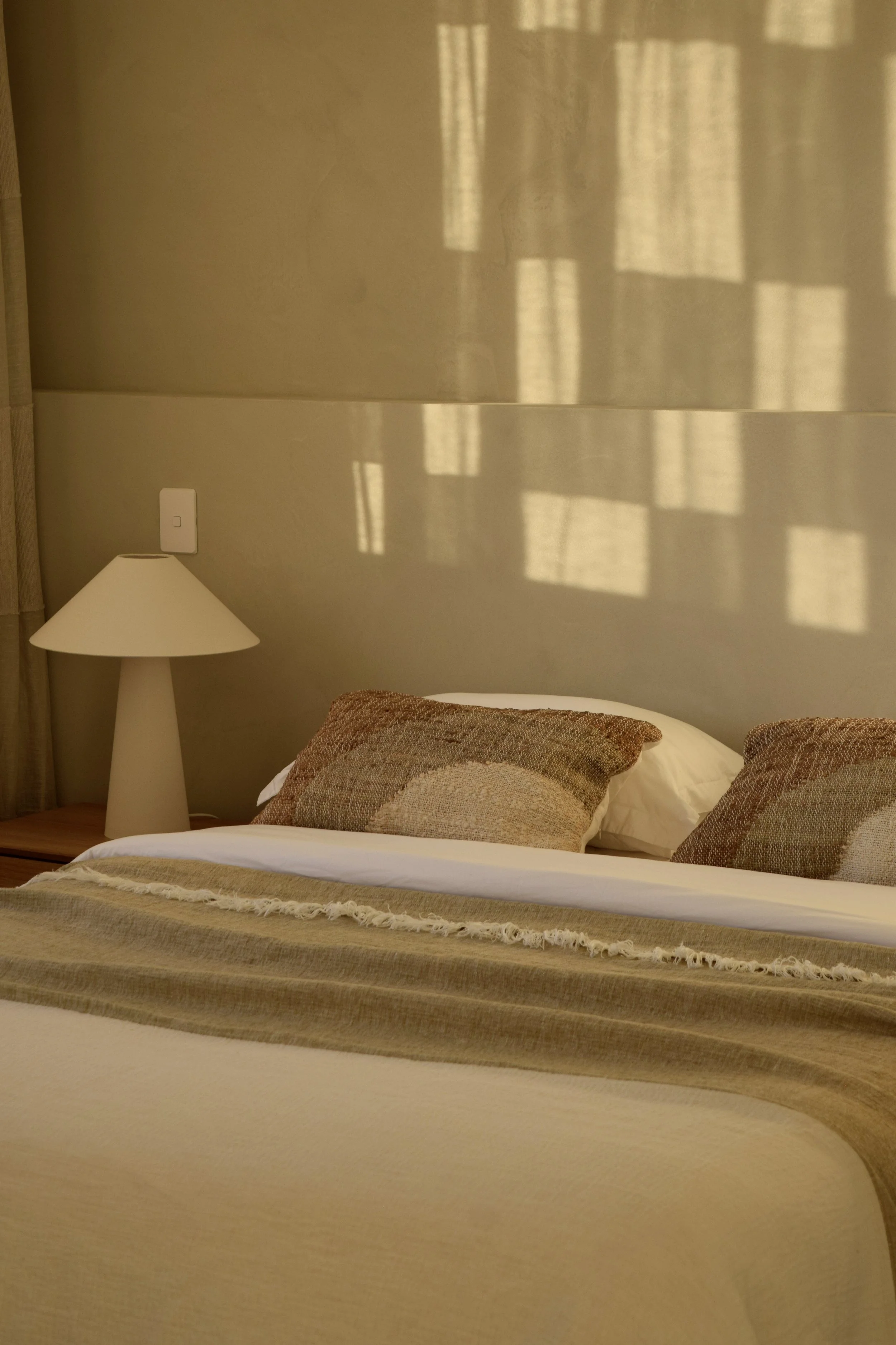A neatly made bed with decorative pillows and a beige blanket, a white bedside lamp, and a beige wall with shadows from window blinds.