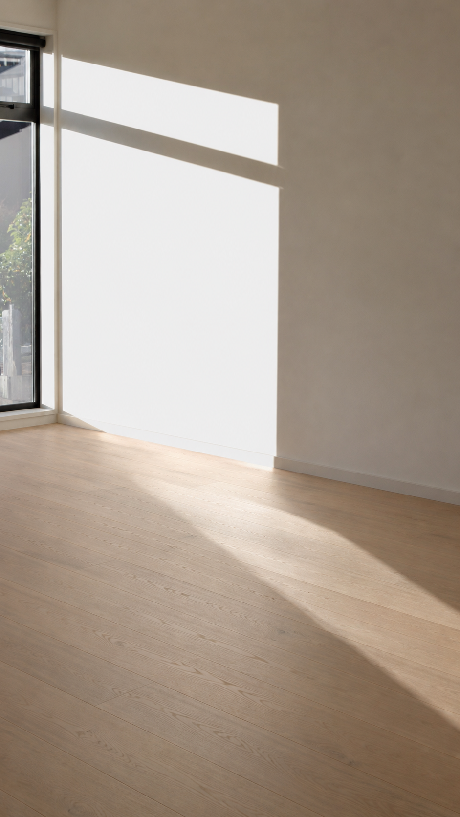 Empty room with wooden flooring and a large window casting sunlight and shadows on the wall and floor.