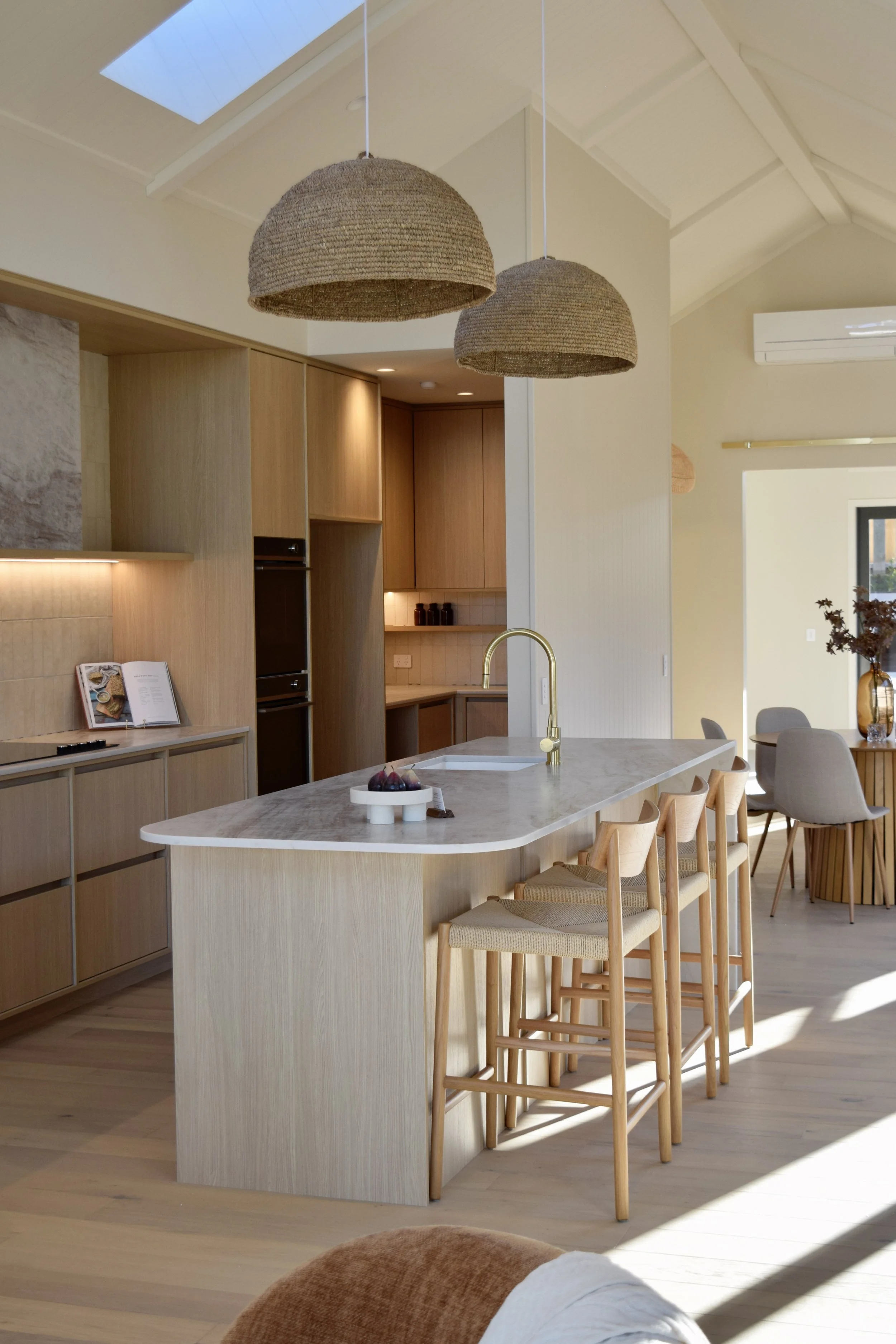Modern kitchen and dining area with a large island, natural wood finishes, hanging woven pendant lights, and a high ceiling with skylights.