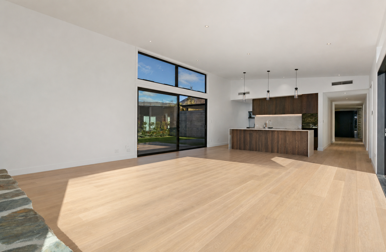 Empty modern living area with hardwood floors, large sliding glass doors, and an open kitchen with hanging pendant lights.